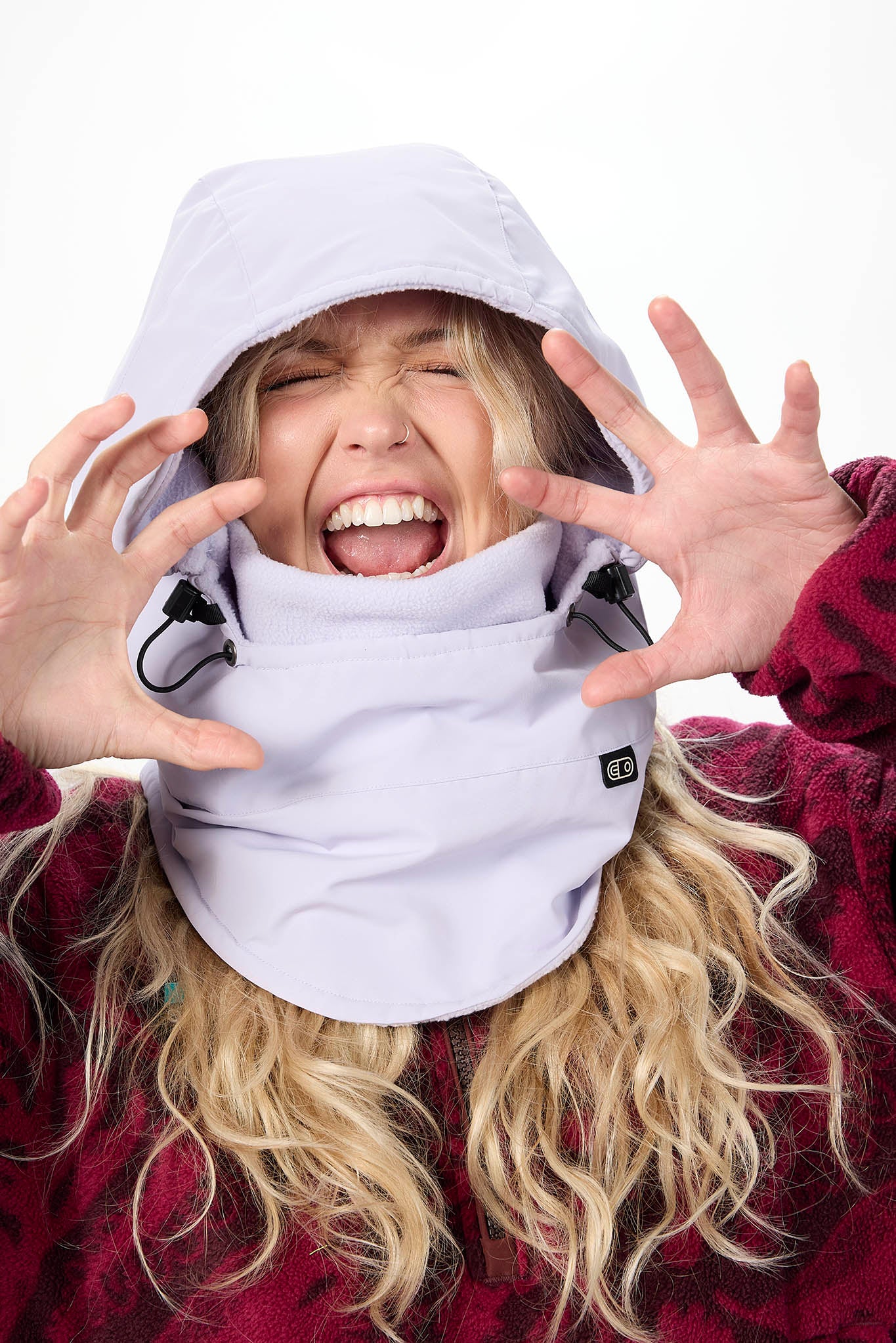 A woman wearing a light lavender heated snow cap and matching neck gaiter, with her hands raised in front of her face against a white background.