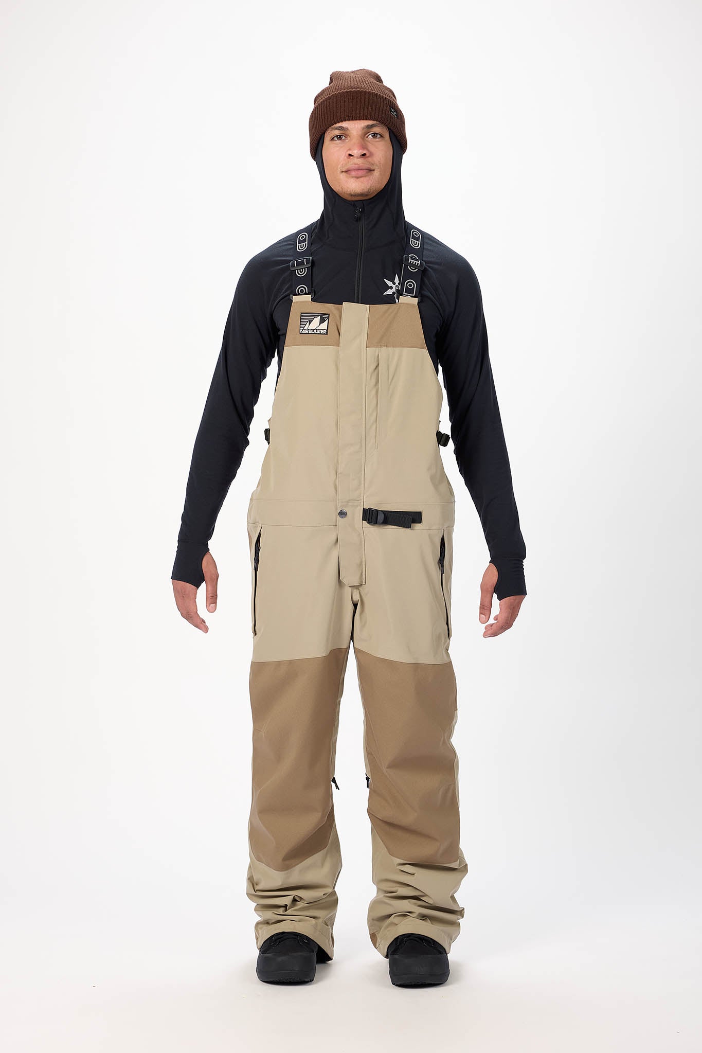 Person wearing a black long-sleeve shirt, brown beanie, and beige bib overalls on a white background