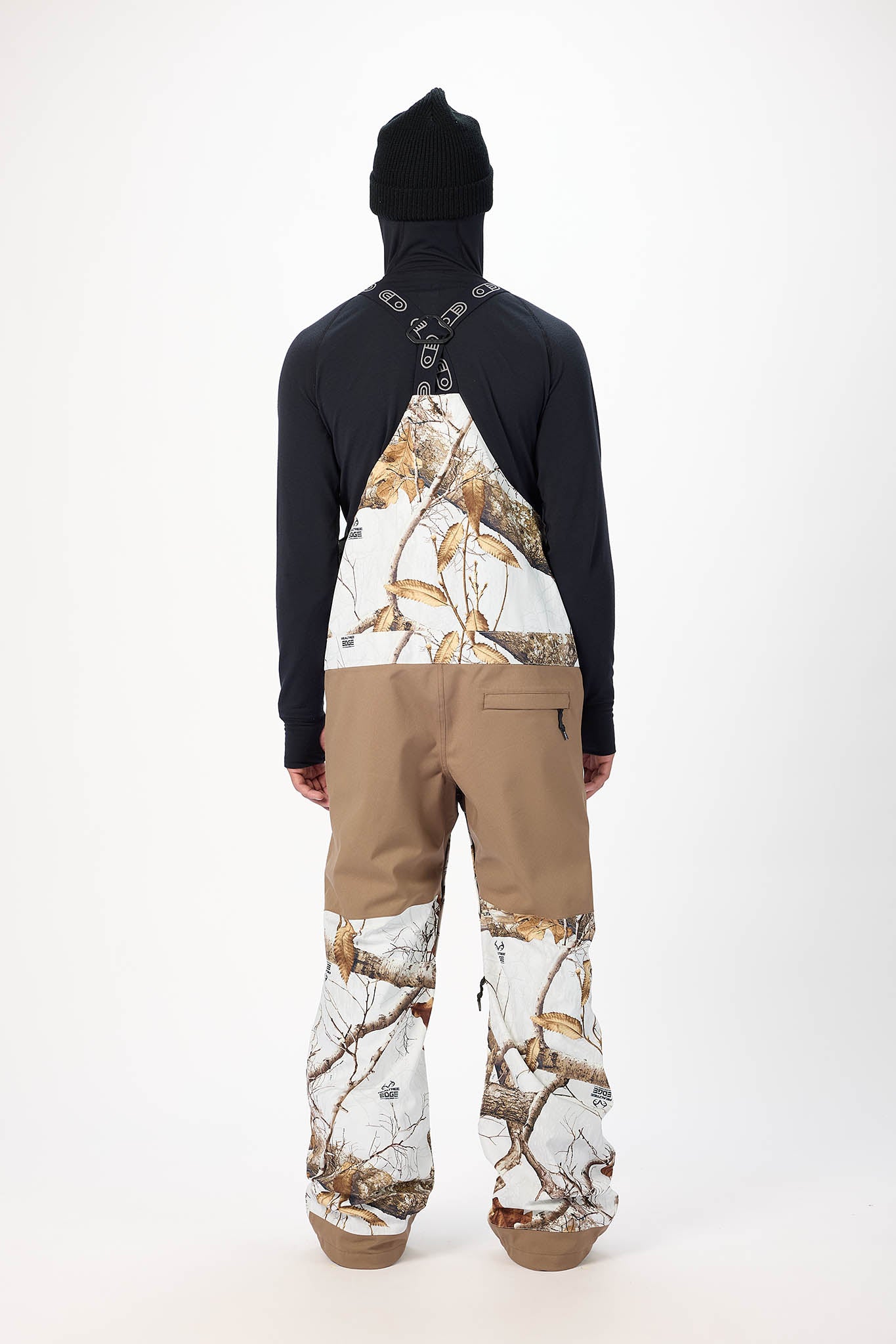 Person wearing a black jacket with a camouflage pattern on the back, beige pants, and a black beanie on a white background.