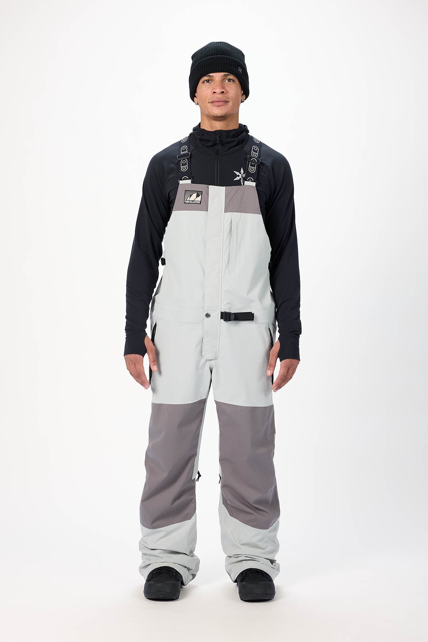 Person wearing a black long-sleeve shirt, gray and white bib overalls, and black beanie on a white background