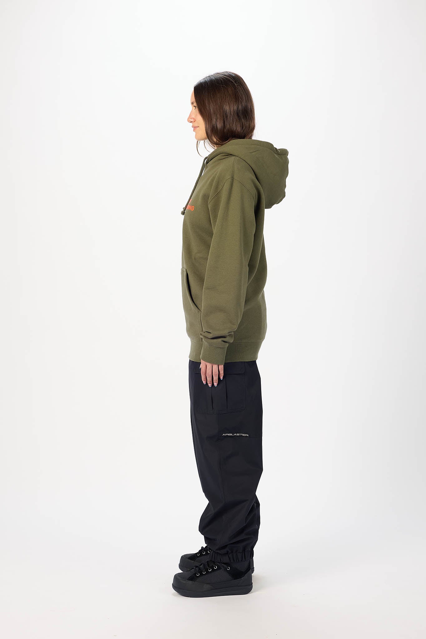 A model shown from the side profile wearing an olive green hooded sweatshirt, black pants, and black sneakers, standing against a plain white studio background.