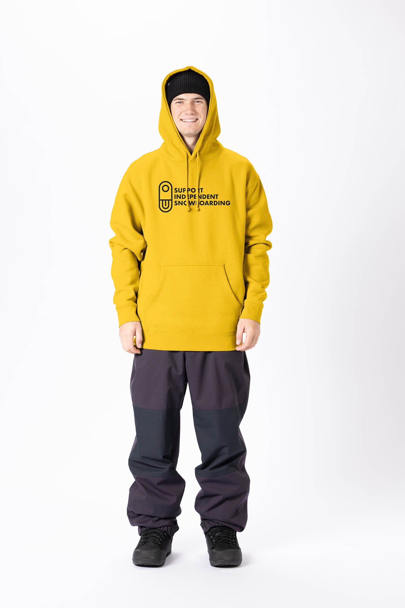 A model stands against a plain white studio background wearing an yellow pullover hoodie with black graphic on the chest, paired with black pants and black sneakers.