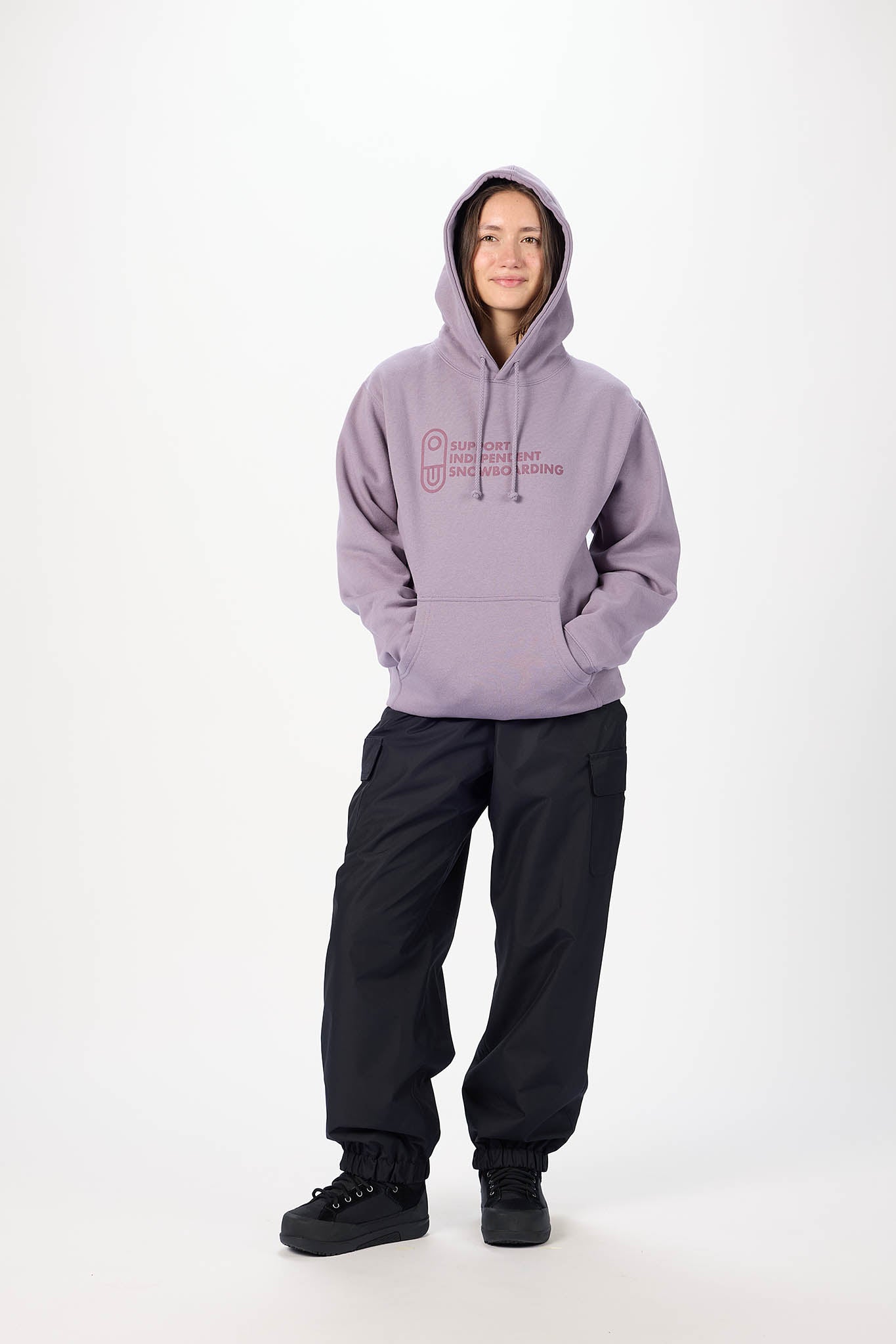 A model wearing a lavender-purple pullover hoodie with a pink graphic on the chest, paired with black pants and black boots, standing against a plain white studio background.