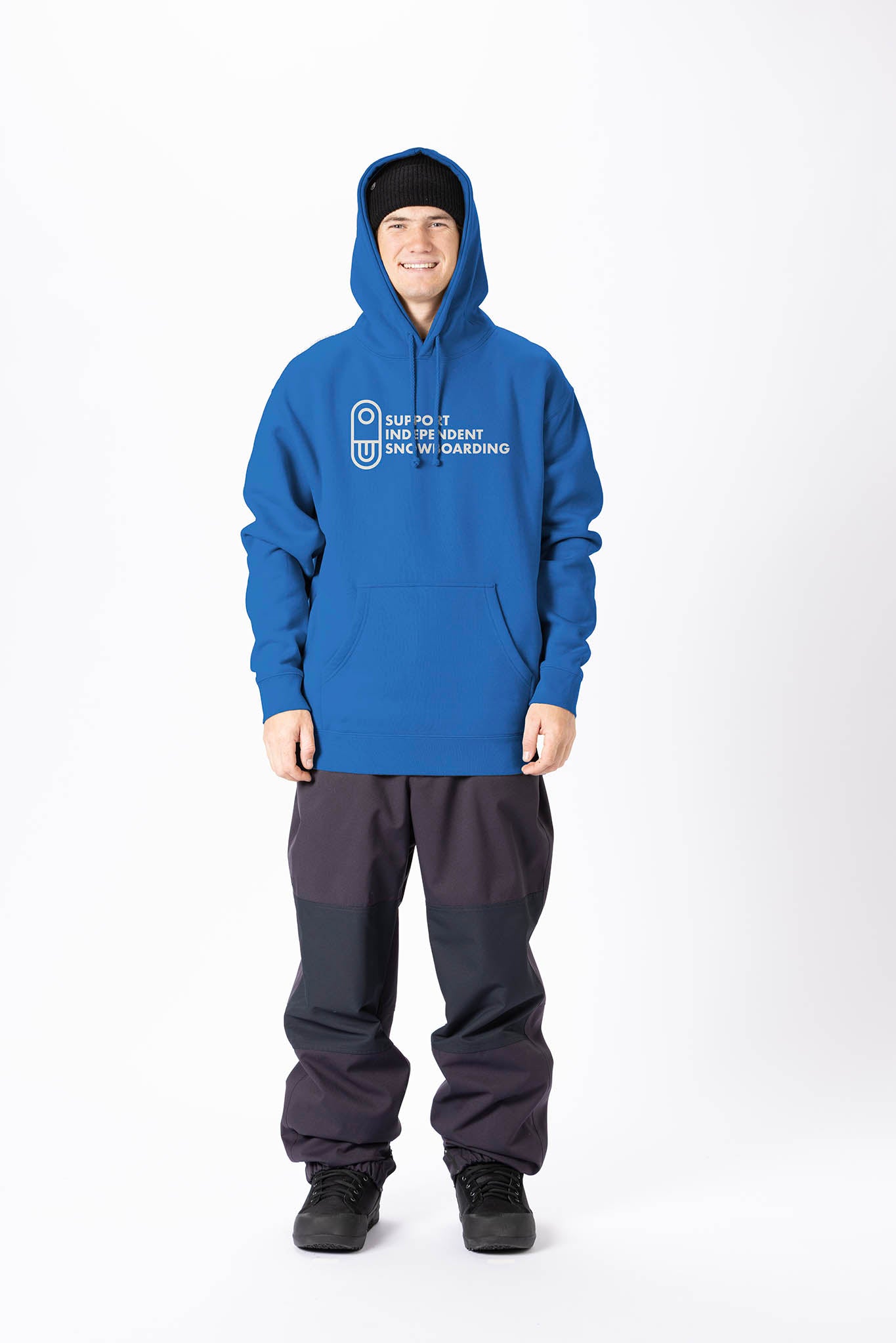 A man wearing a blue pullover hoodie with a white printed graphic on the chest, paired with dark gray and black pants, standing against a plain white studio background. 