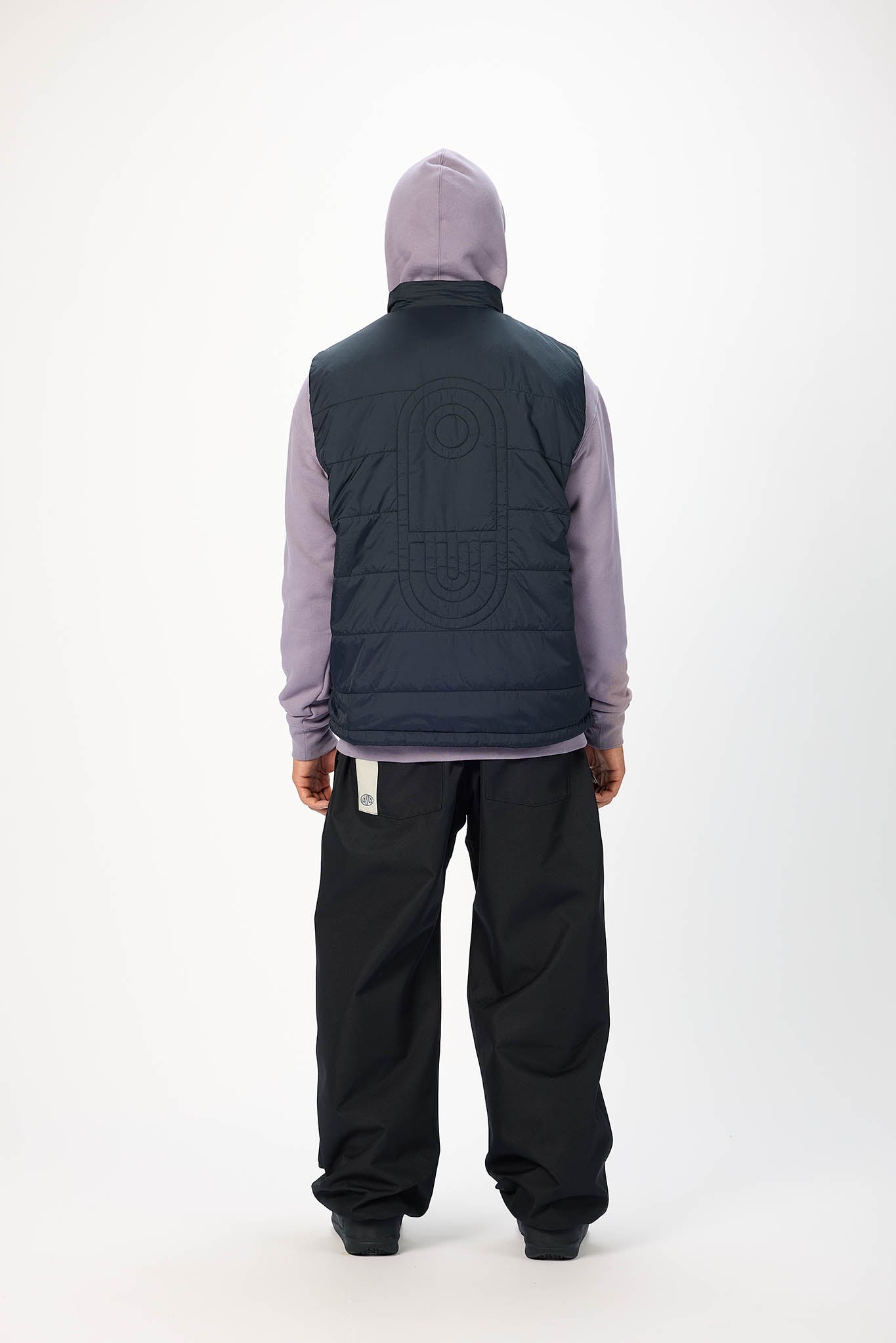 A person shown from the back wearing a dark navy quilted puffer vest over a light purple hoodie and black pants, standing against a plain white studio background.