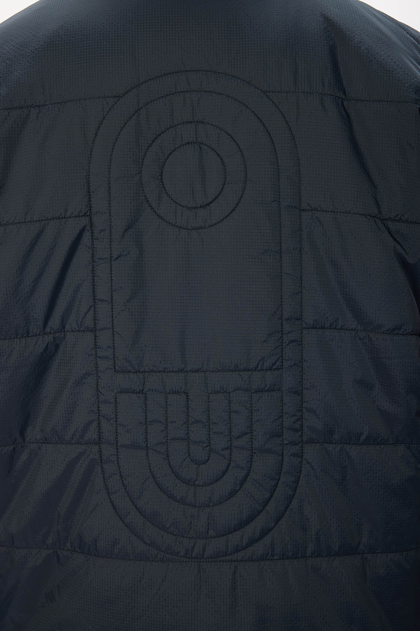 Close-up of a dark navy quilted puffer jacket showing a large tonal embroidered geometric logo on the back.