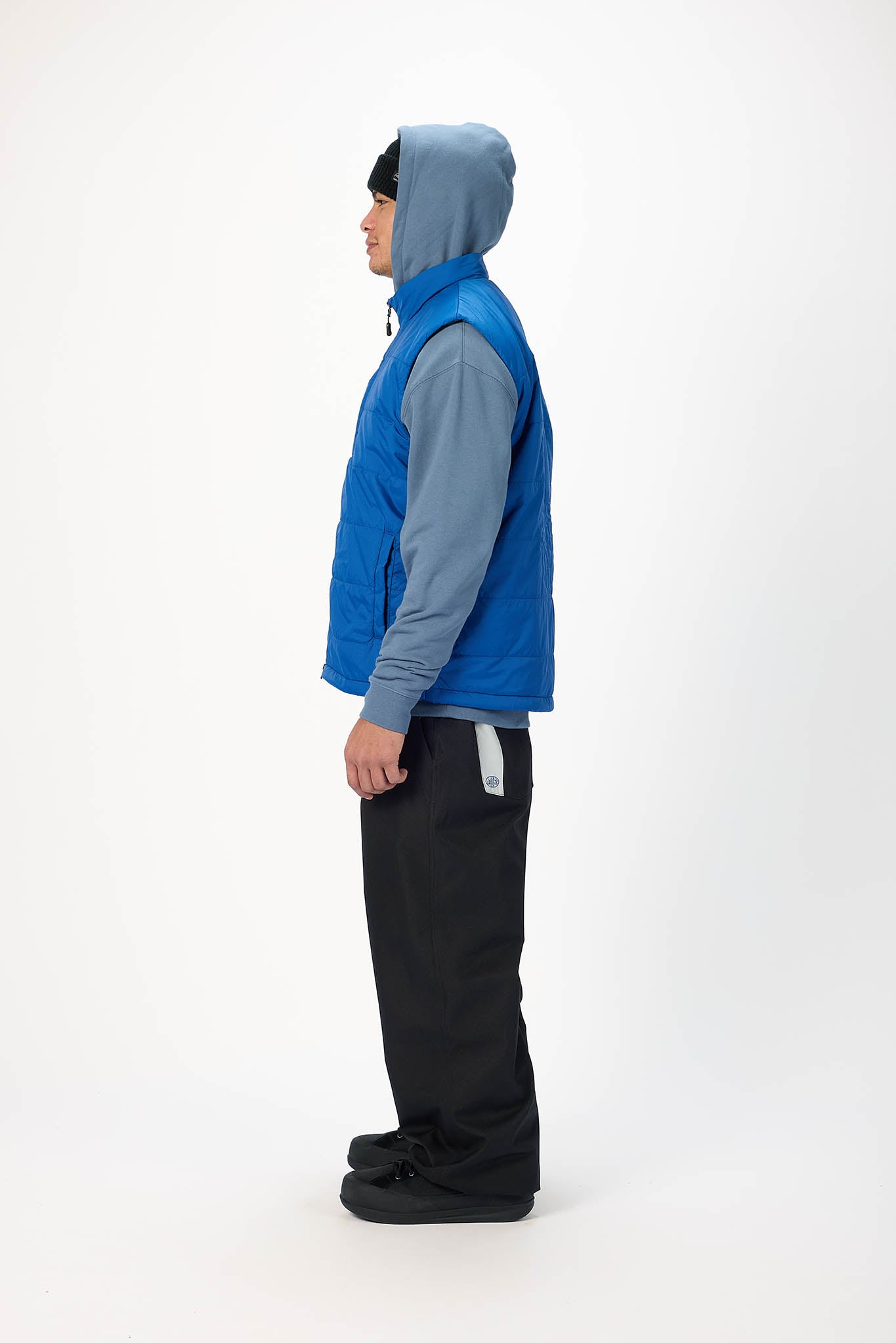 A man shown in profile wearing a bright blue hooded puffer vest over a heather gray long-sleeve shirt, black pants, and black shoes, standing against a plain white studio background.