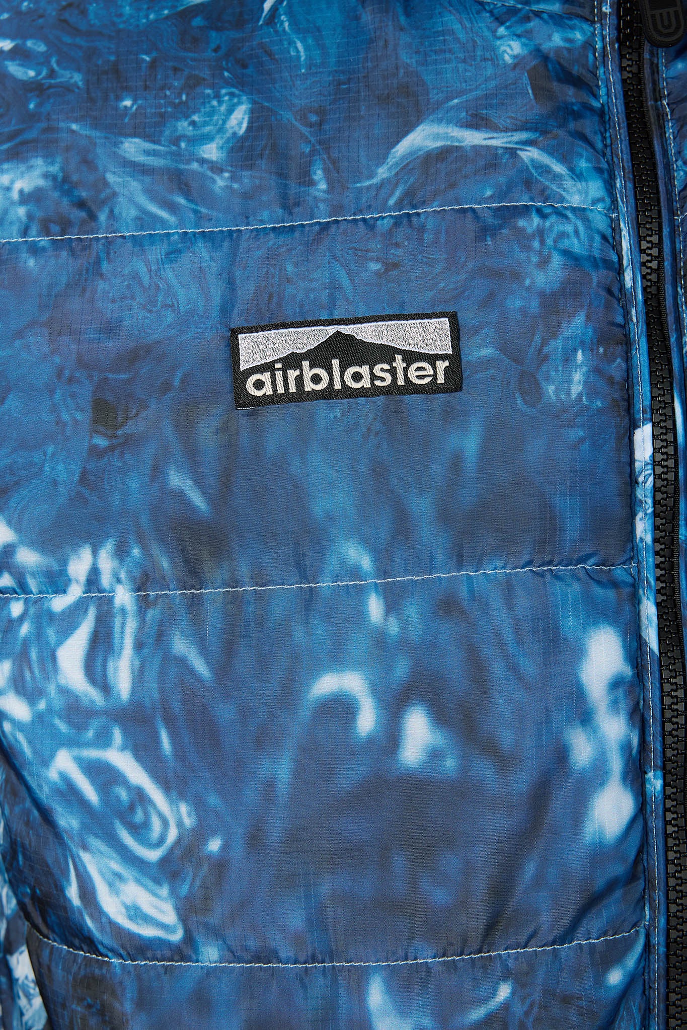 Close-up of a blue quilted puffer jacket with a glossy, marbled pattern and a black zipper. A black rectangular patch on the chest features the white 'airblaster' brand name.