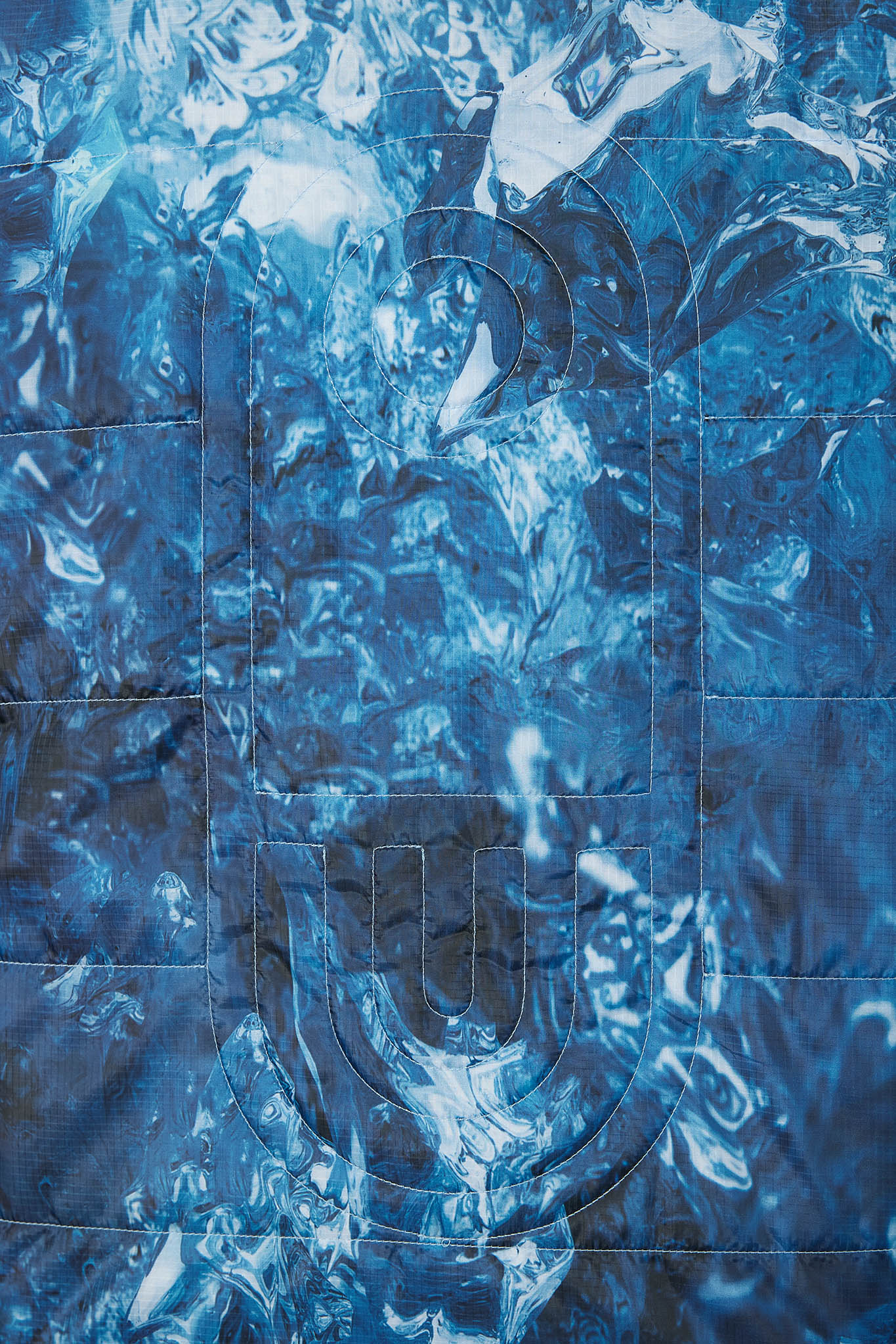 A close-up of a blue quilted fabric featuring a marbled, liquid-like pattern with stitched geometric and curved outlines.