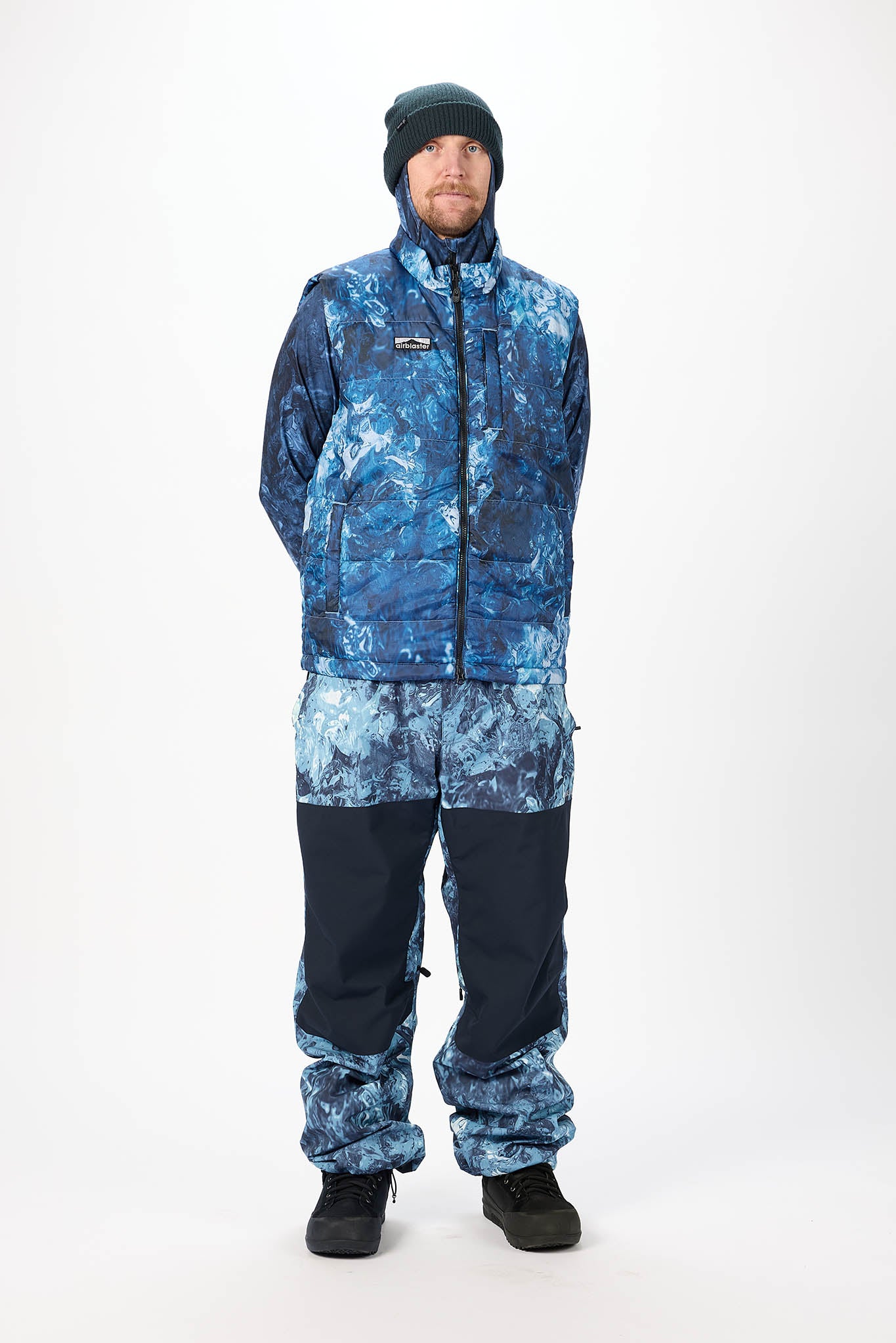 A man modeling a blue and navy winter snowsuit with a blue and white abstract pattern, paired with a dark beanie and black snow boots, against a plain white studio background.