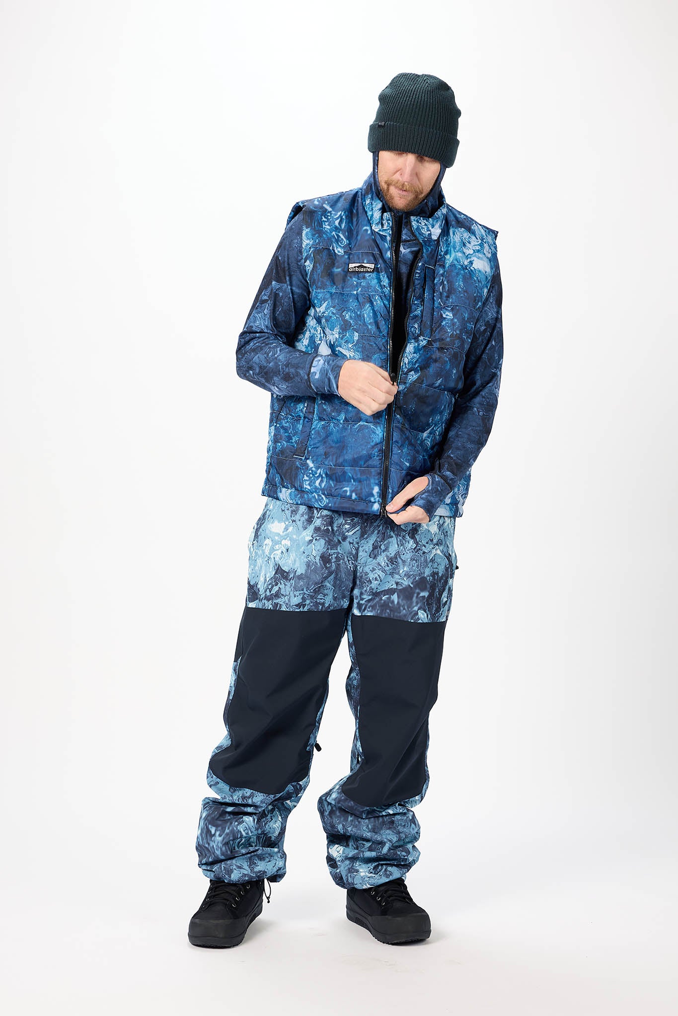 A man modeling a blue and black camouflage snowsuit with a black beanie and black snowboard boots, standing against a white studio background.