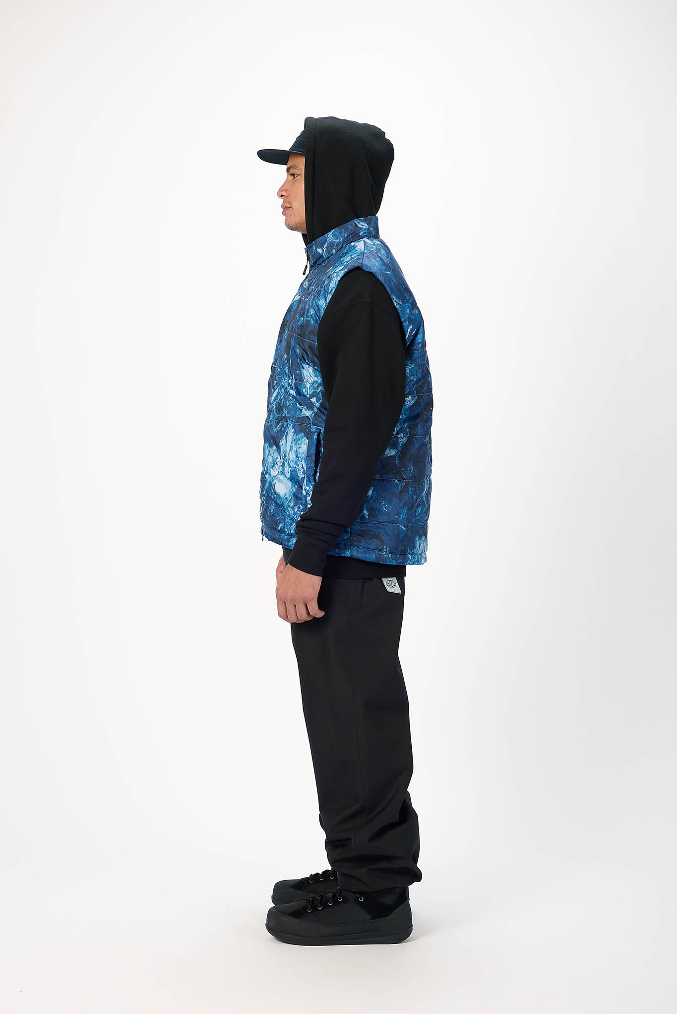 A person shown in profile wearing a blue and black patterned puffer vest over a black hoodie, black pants, and black shoes, standing against a plain light gray studio background.