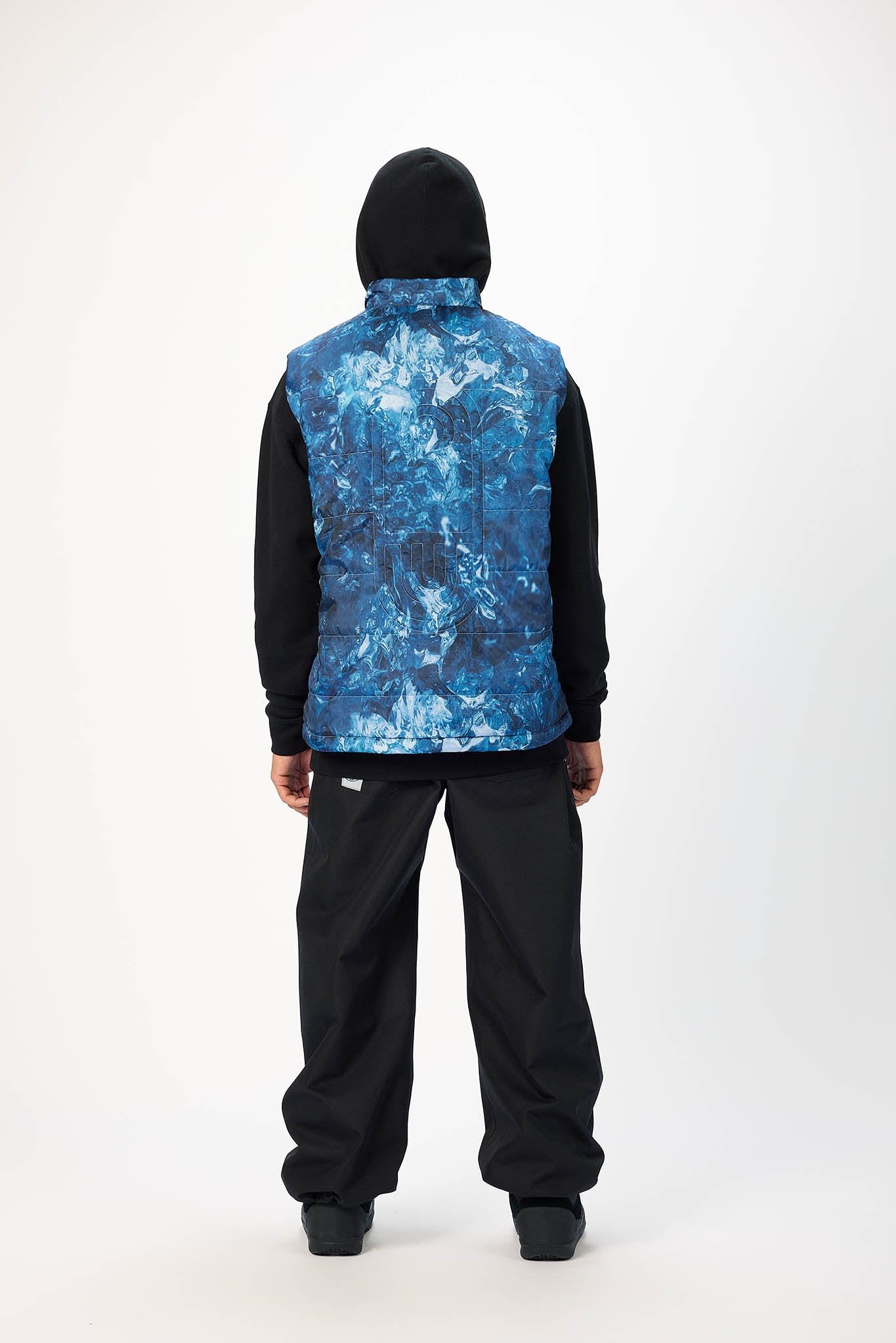 A person shown from the back wearing a black hooded jacket with a vibrant blue and white abstract patterned vest, paired with black pants, against a plain white studio background.