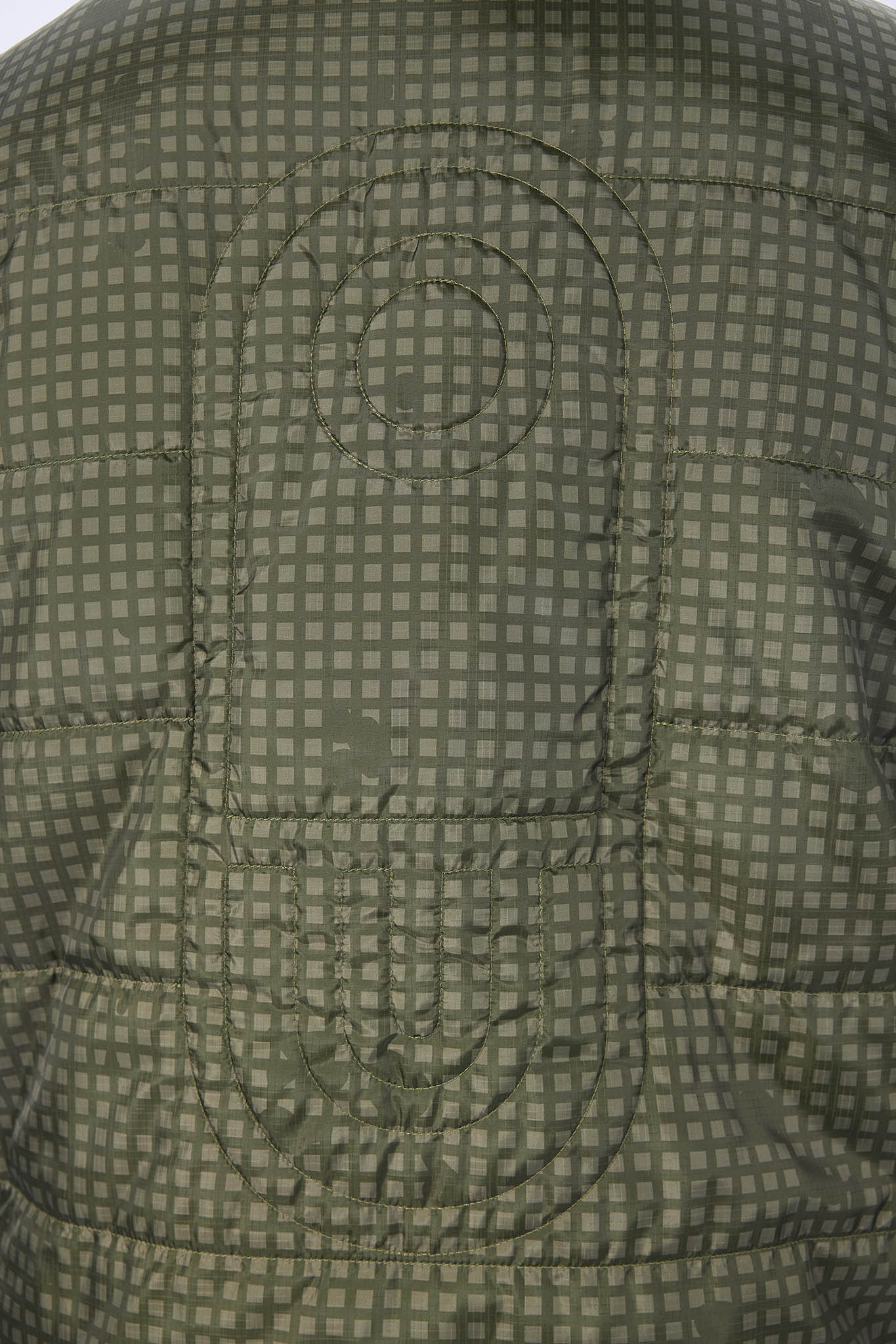Close-up of a quilted jacket or puffer vest showing a small checkered pattern in olive green and gray, with a circular stitched emblem on the chest.