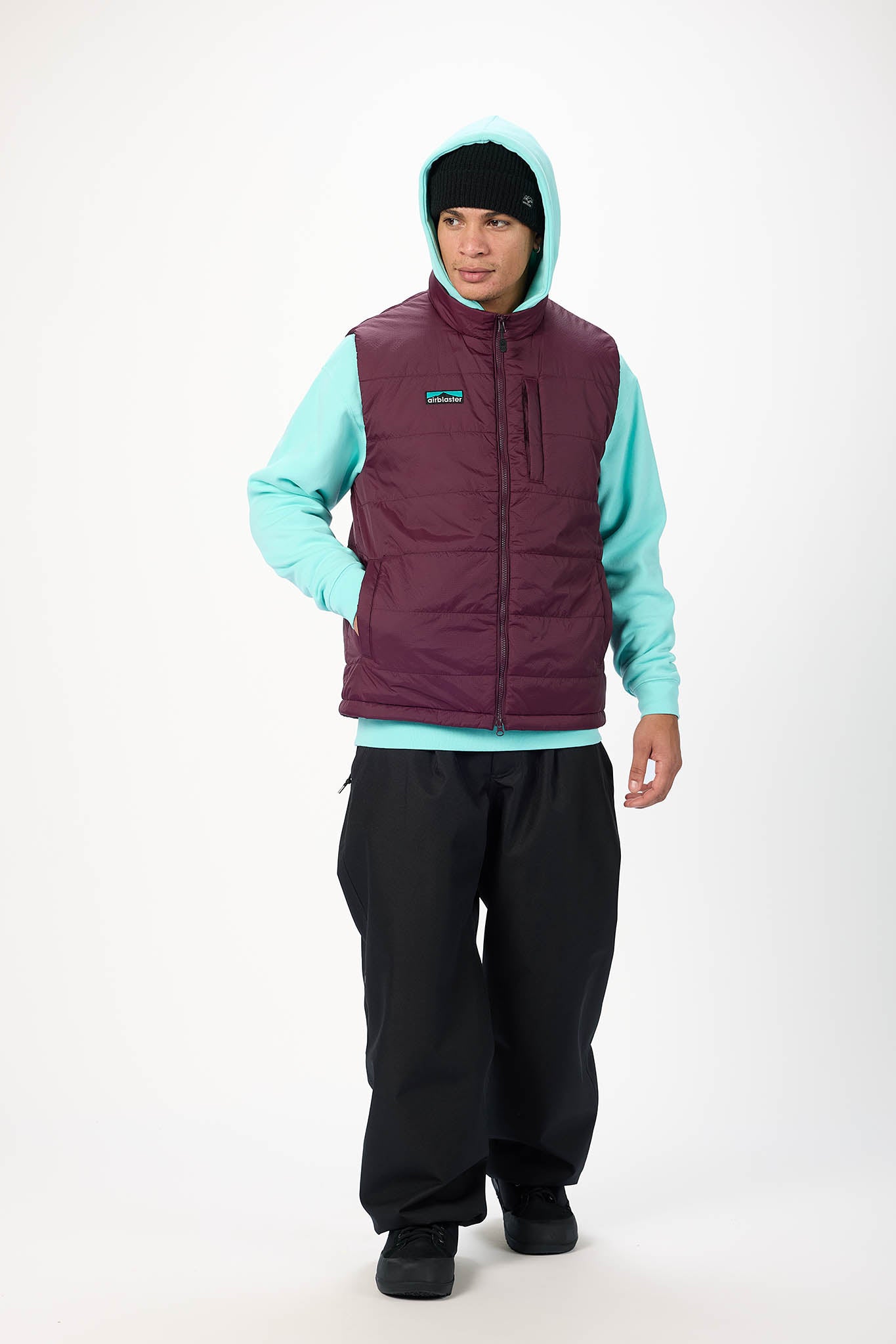 A model wearing a Patagonia insulated sleeveless vest in burgundy, layered over a light aqua hoodie, black pants, and a black beanie, standing against a plain white studio background.