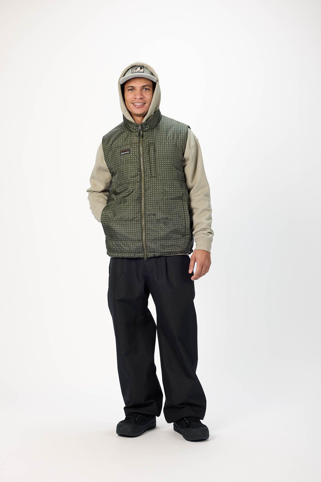 A man wearing a green and black patterned zip-up vest over a beige and gray hooded sweatshirt, black pants, and black shoes, standing against a plain white studio background.