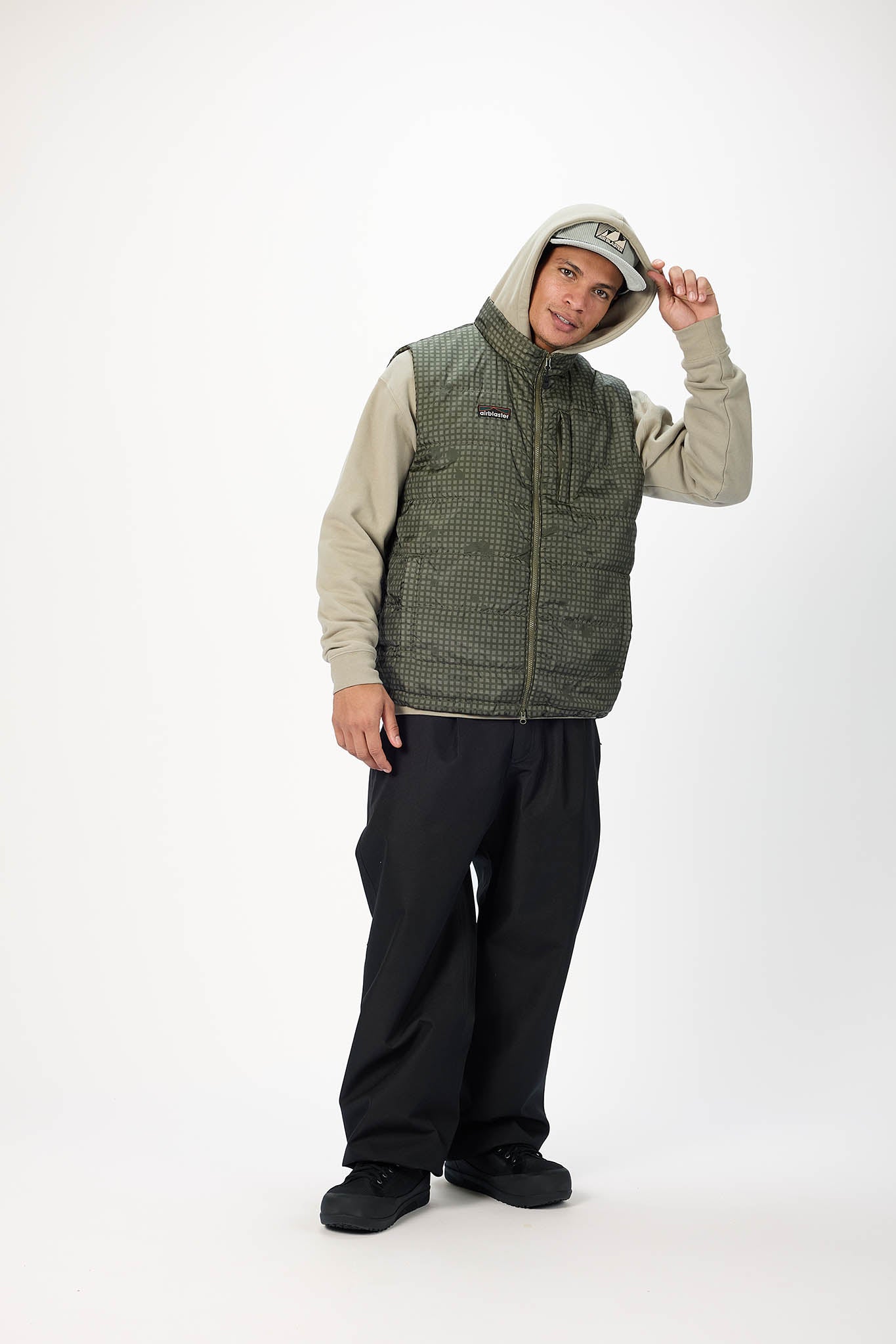 A man modeling a Patagonia zip-up vest over a beige hoodie, black pants, and black boots against a plain white studio background.