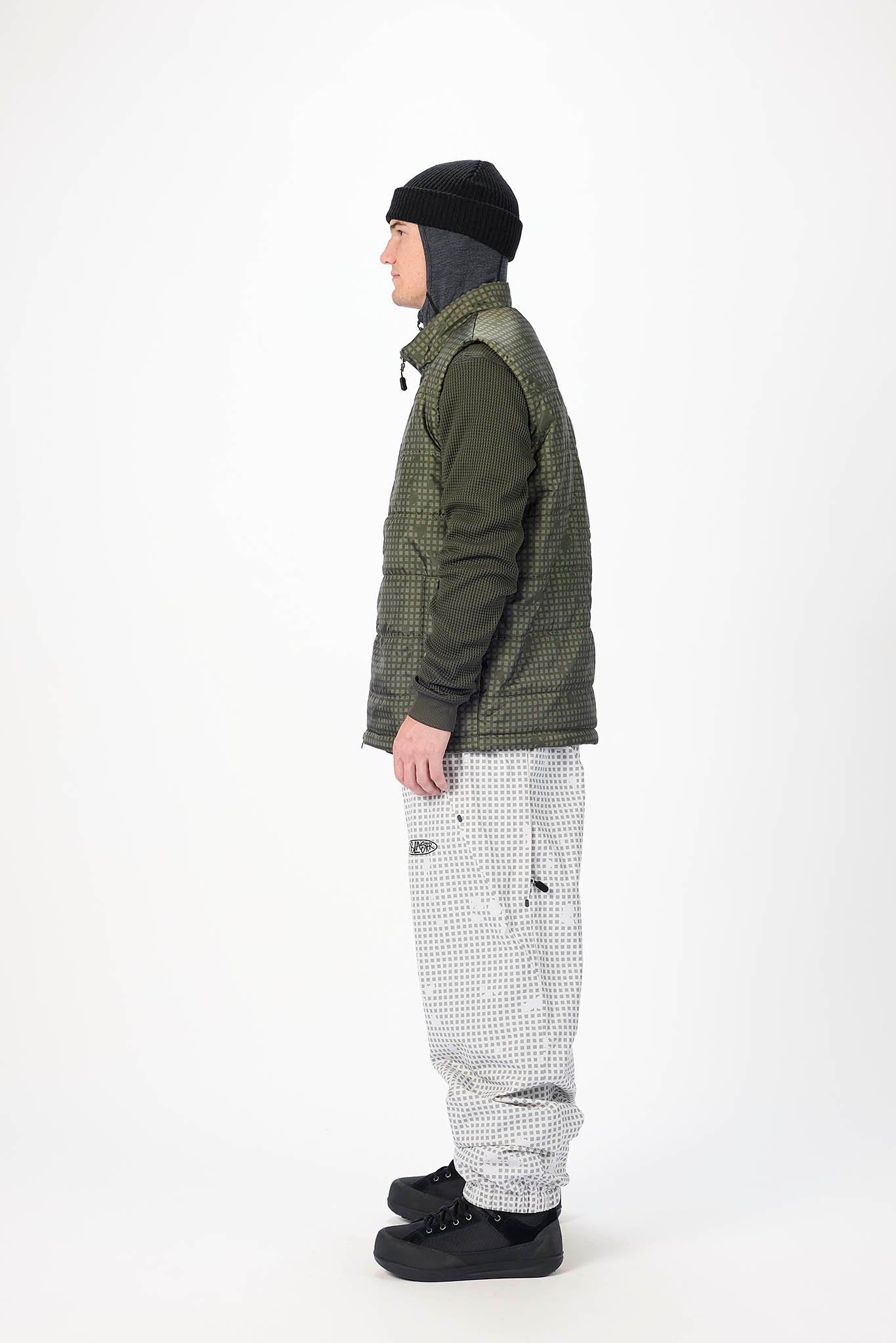 A man shown in profile wearing a dark olive green quilted puffer vest over a dark long-sleeve shirt, paired with light gray and black checkered snow pants, a black beanie, and black snow boots, against a plain white studio background.