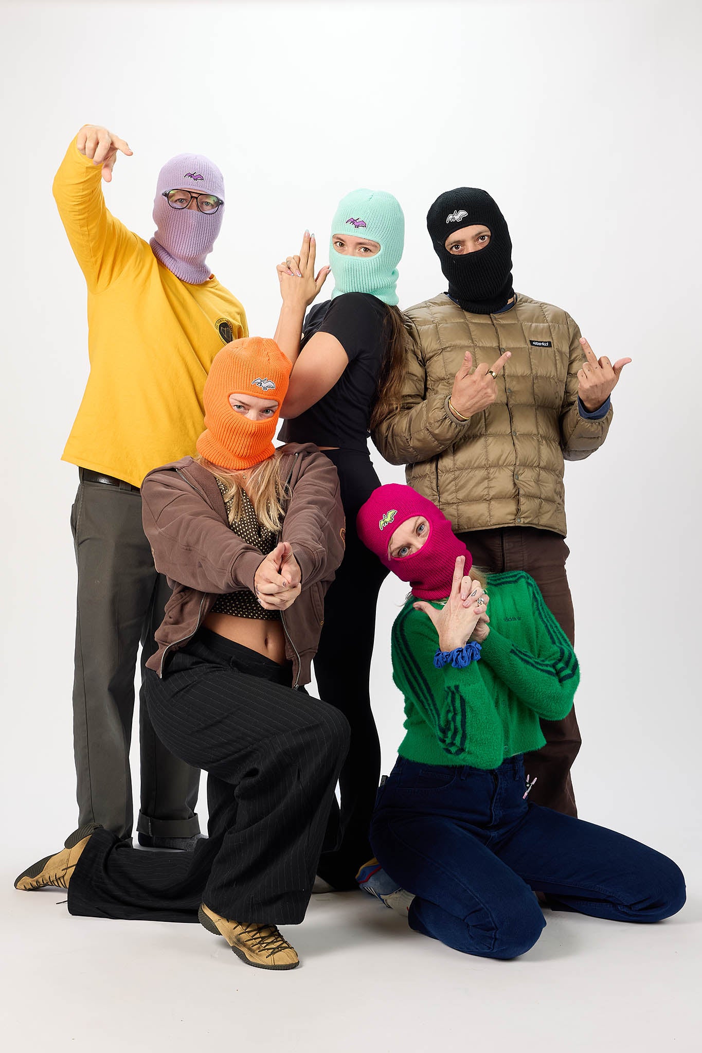 Five people posing together in a studio setting, each wearing a different colored ski mask (lavender, mint, black, orange, and magenta) with various streetwear outfits.