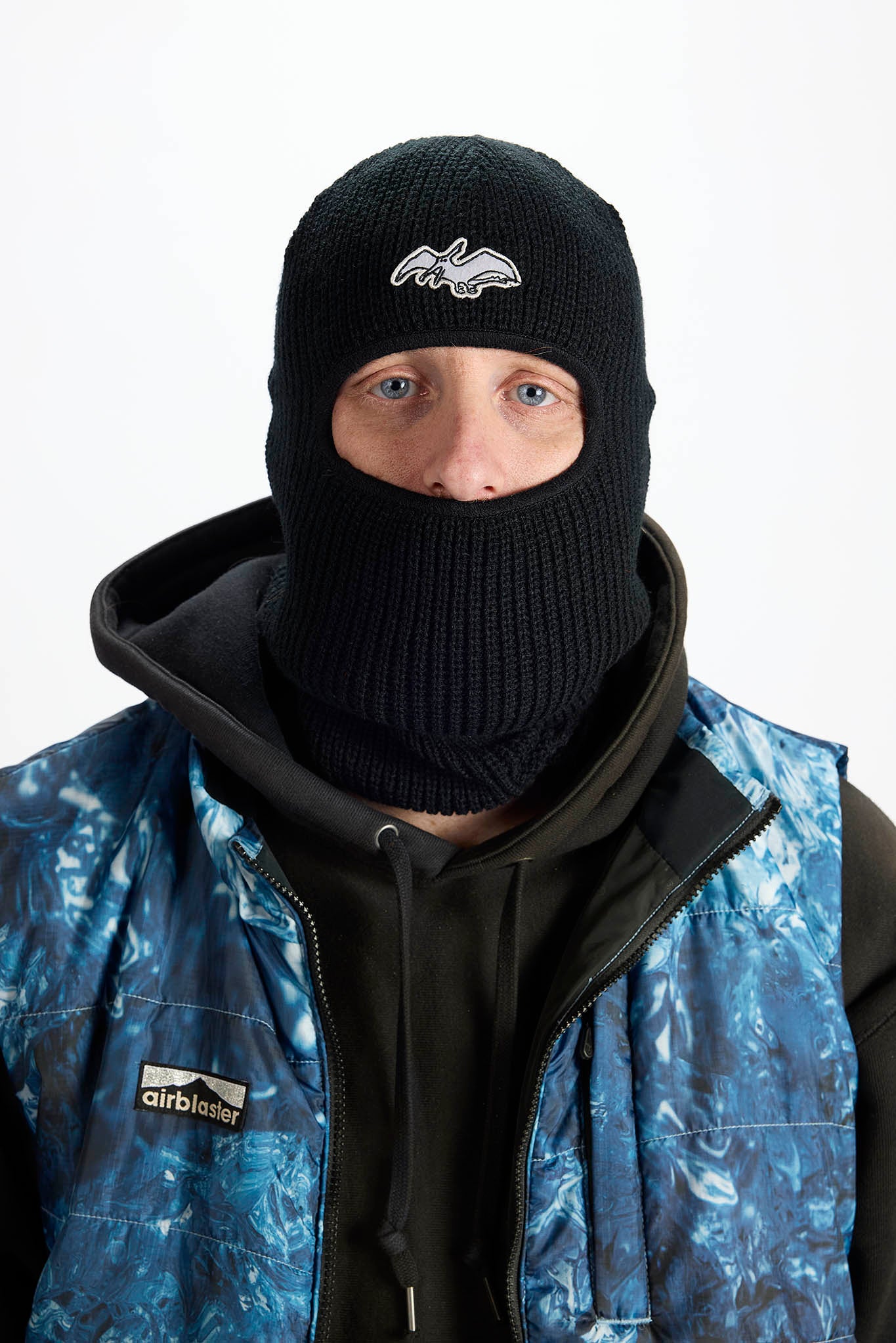 A person wearing a black knit ski balaclava with a white logo on the front, layered under a blue and black insulated vest.