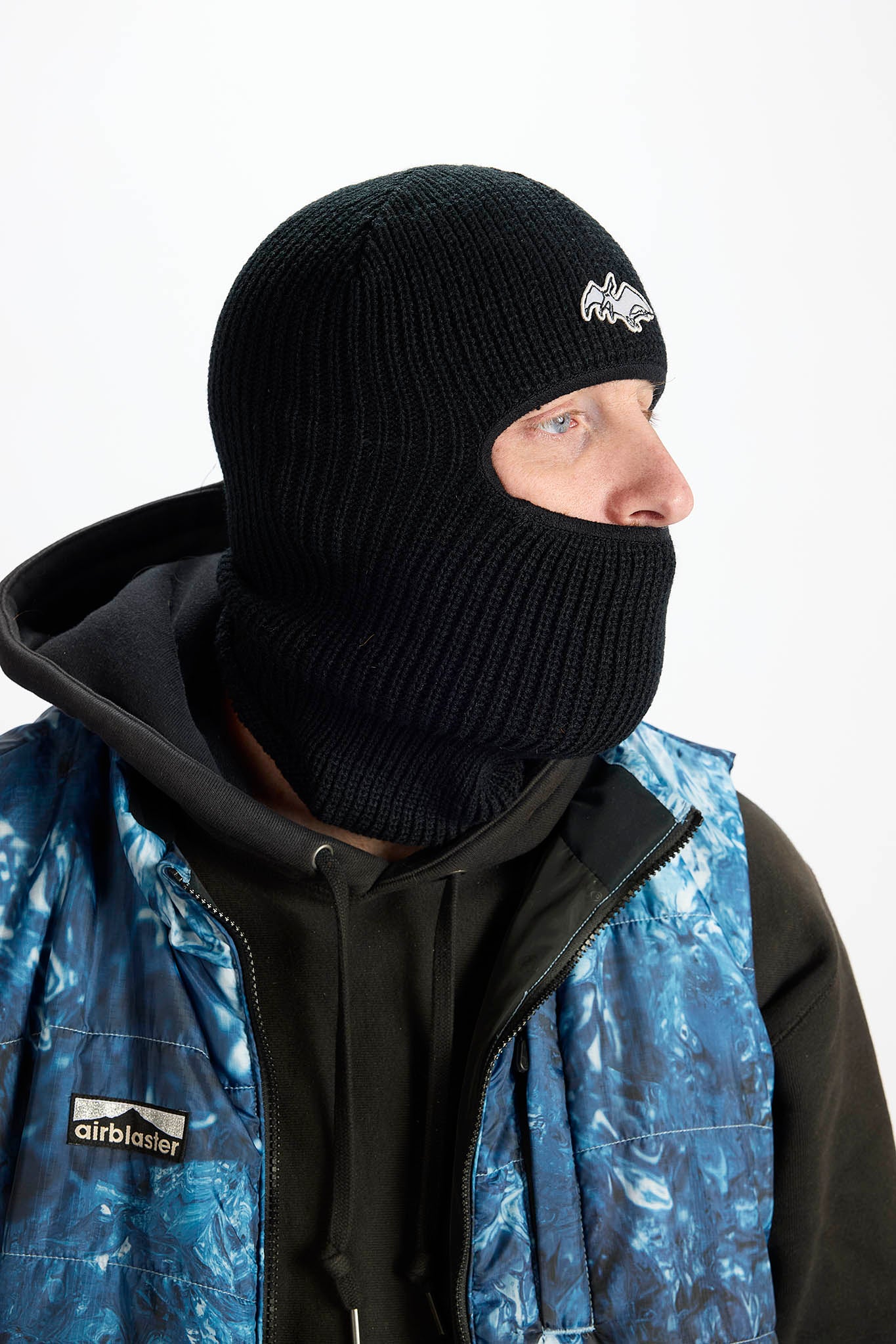 A man wearing a black knit balaclava mask with a small white logo on the side, paired with a blue and black Airblaster jacket against a plain white background.