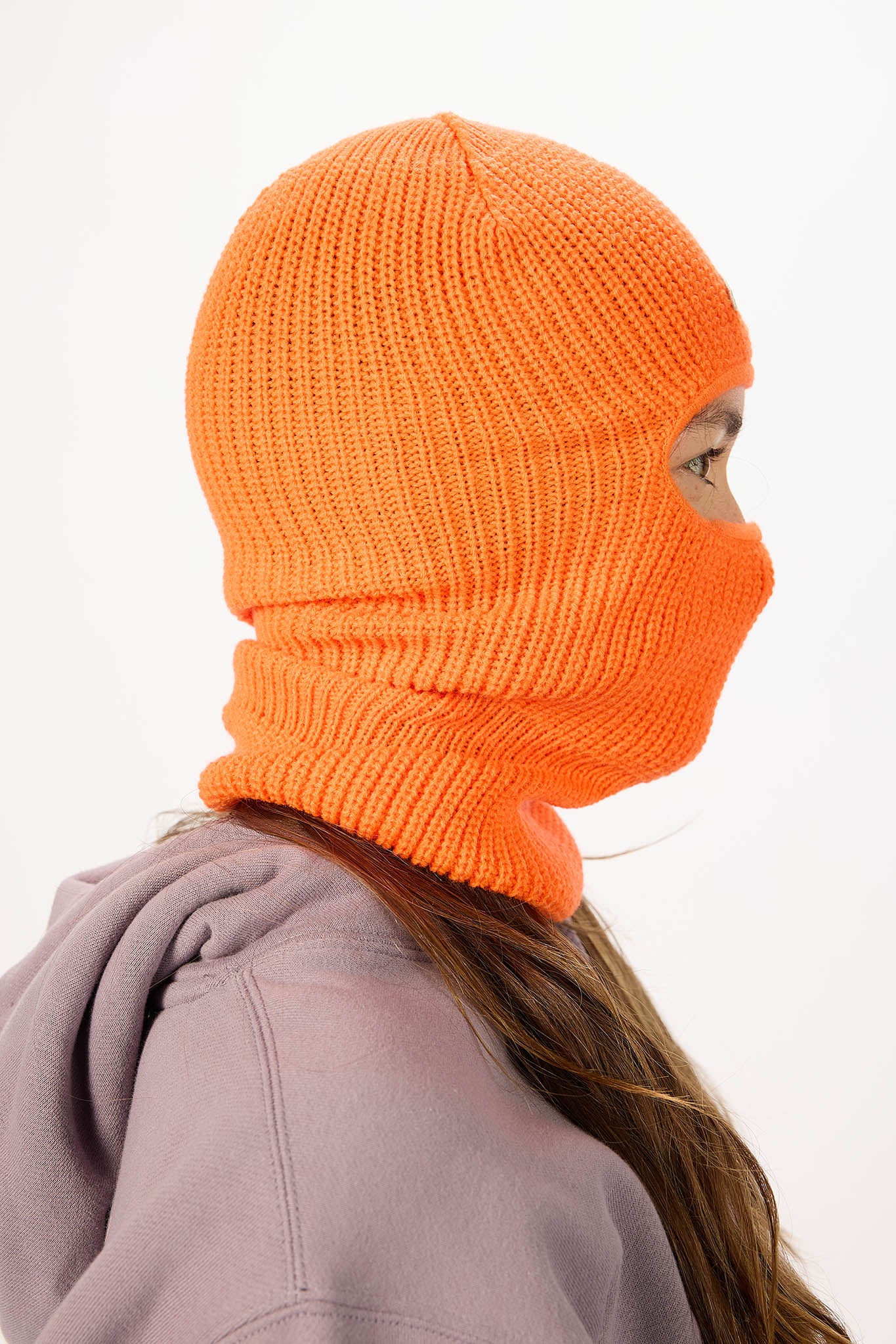 A bright orange ribbed knit balaclava mask worn by a model, shown in profile against a plain white background.