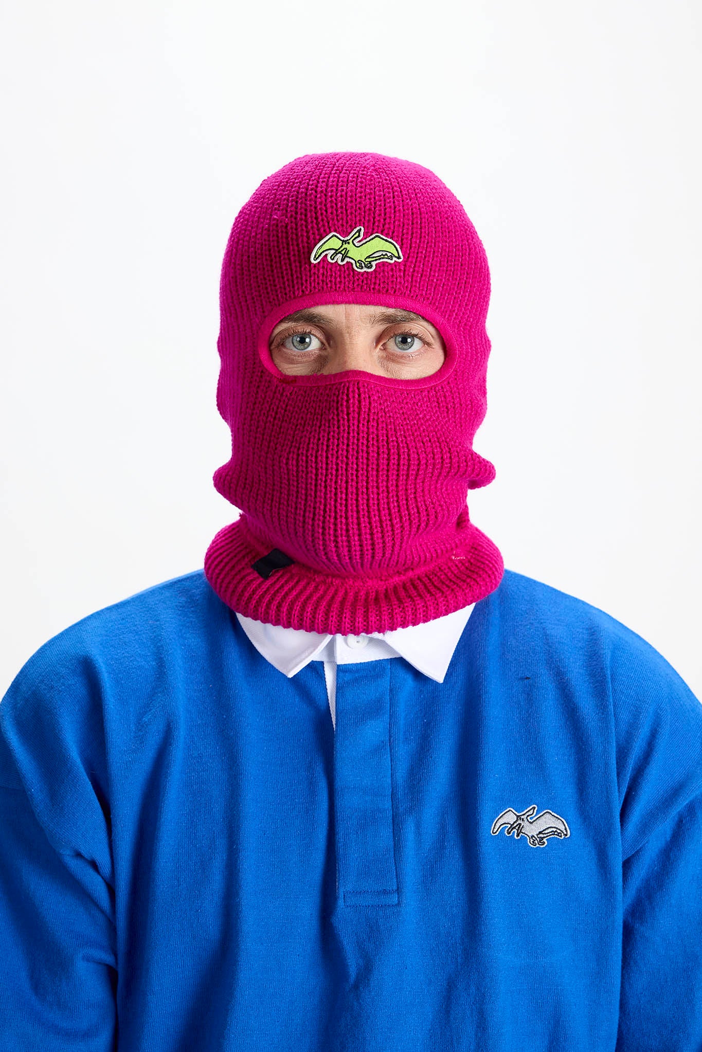 A person wearing a bright pink knit balaclava mask with a small embroidered logo on the forehead, paired with a blue polo shirt against a plain white background.