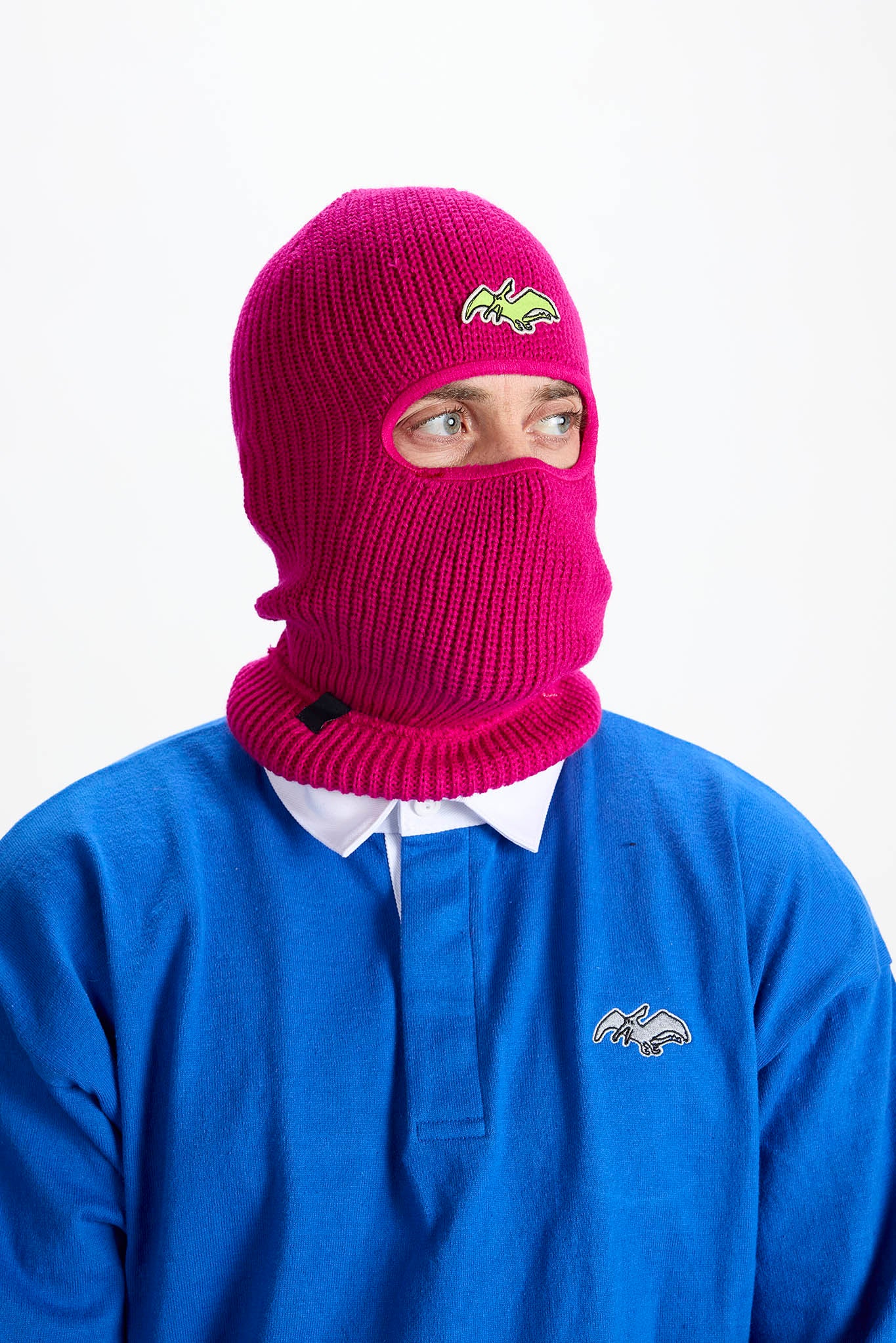 A person wearing a bright magenta knit balaclava mask with a small embroidered logo on the forehead, paired with a blue polo shirt against a plain white background.