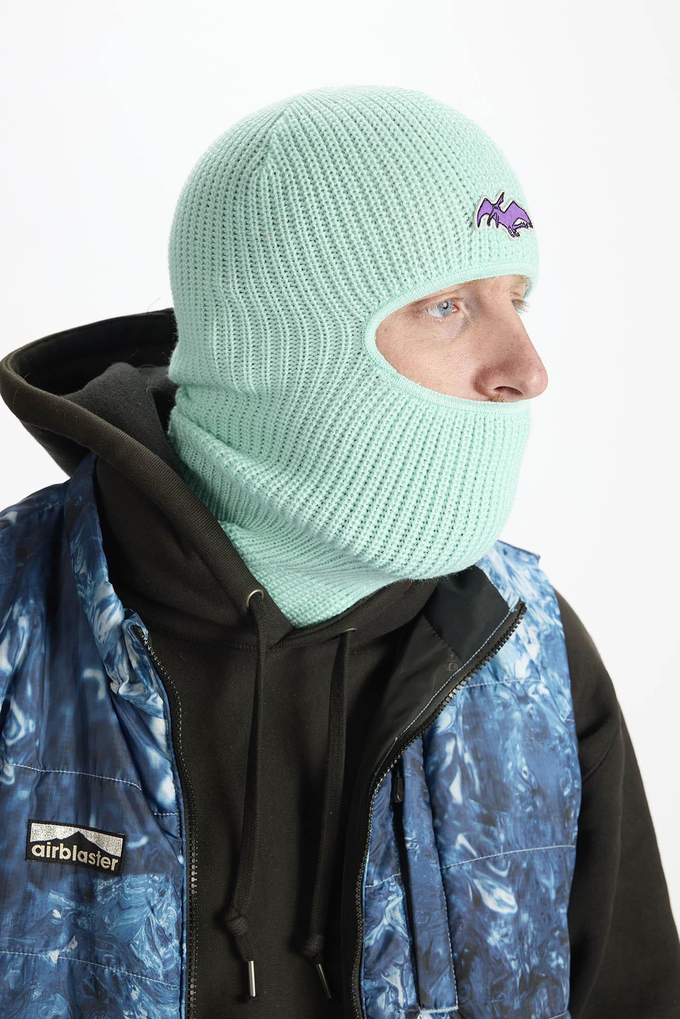 A person wearing a mint green knit balaclava mask with a small purple Airblaster logo, layered over a black hoodie and a blue patterned Airblaster jacket, against a plain white background.