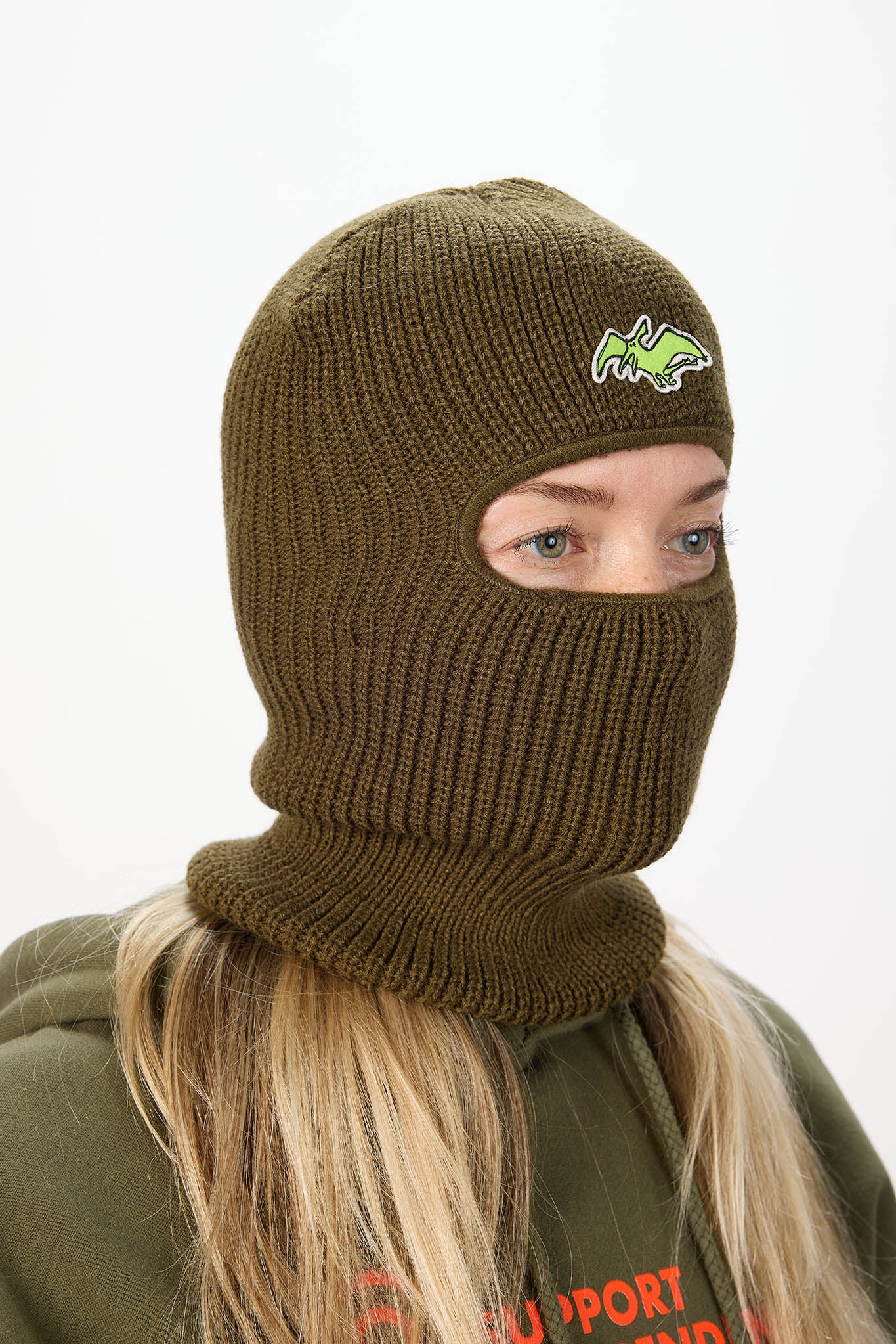 A person wearing an olive green ribbed knit balaclava mask with a small green and yellow embroidered logo on the forehead, shown against a plain light background.