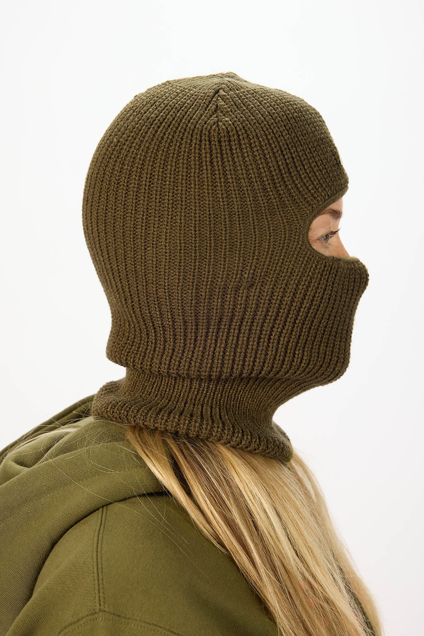 A person shown in side profile wearing an olive green ribbed knit balaclava mask and matching hooded jacket against a plain white background.