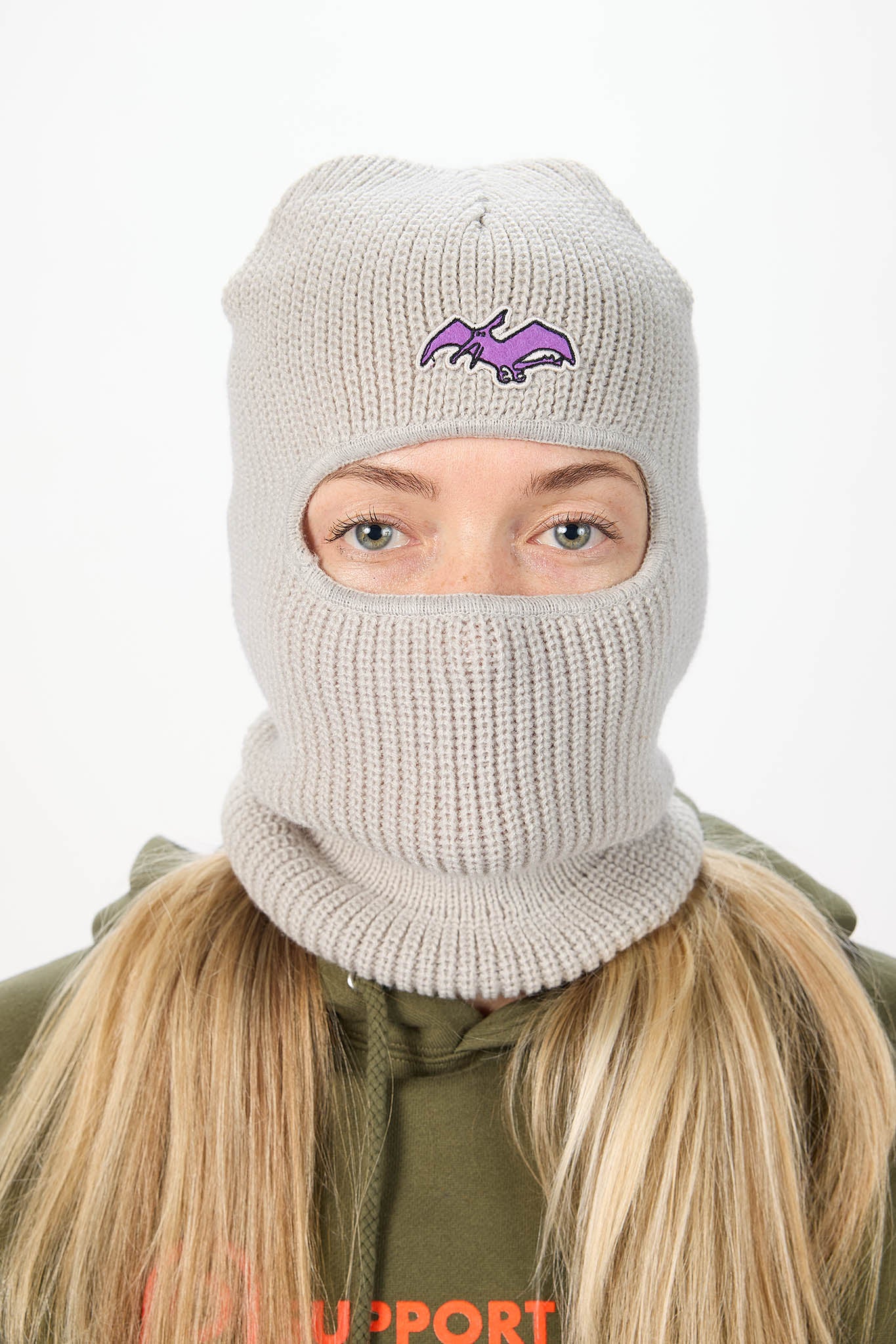 A woman wearing a light gray ribbed knit balaclava mask with a small purple terry logo on the forehead, shown against a plain white background.