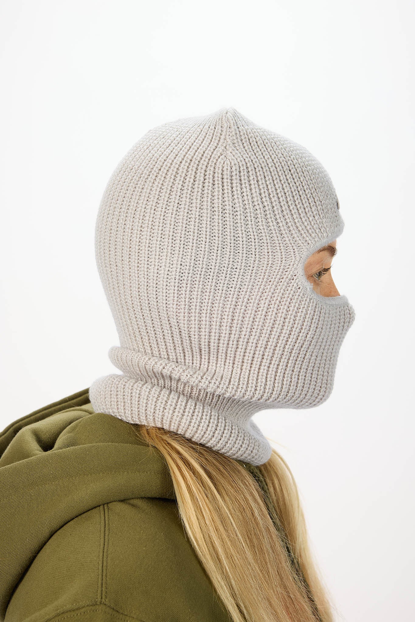 A side-profile view of a person wearing a light gray ribbed knit balaclava mask and an olive green hoodie against a plain white background.