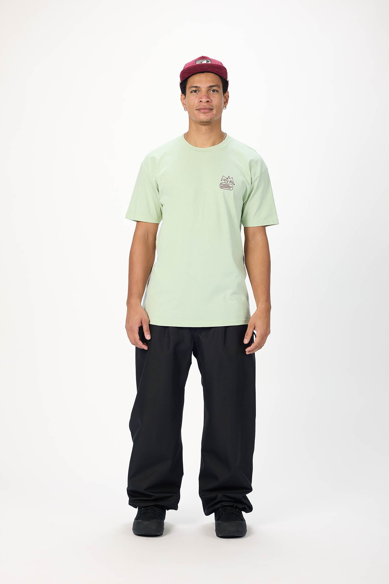 A man modeling a light green crewneck t-shirt with a small graphic on the left chest, paired with black pants and a red baseball cap, standing against a plain white studio background.
