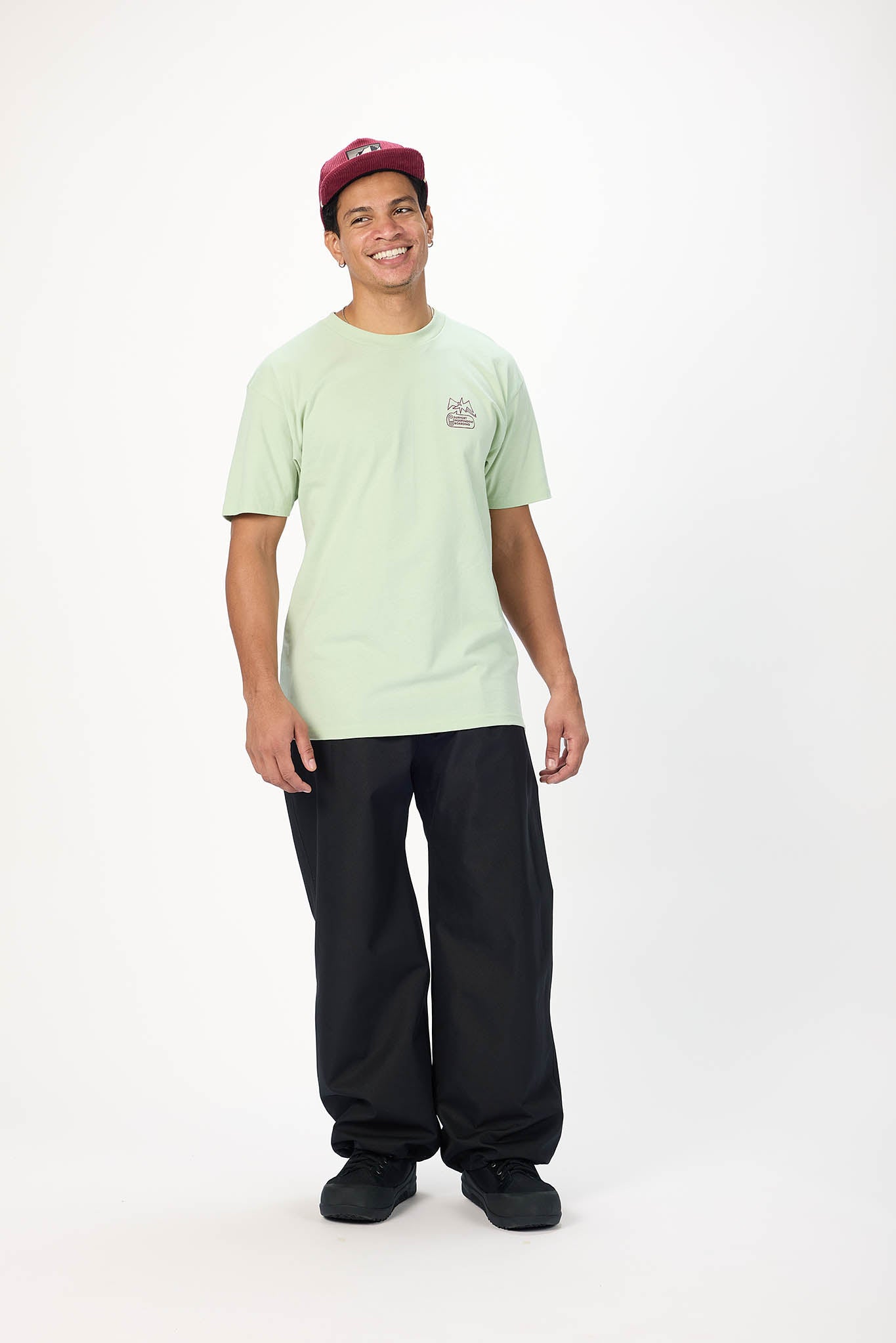 A young man wearing a light sage green t-shirt with a small chest graphic, black pants, and a burgundy baseball cap, standing against a plain white studio background.