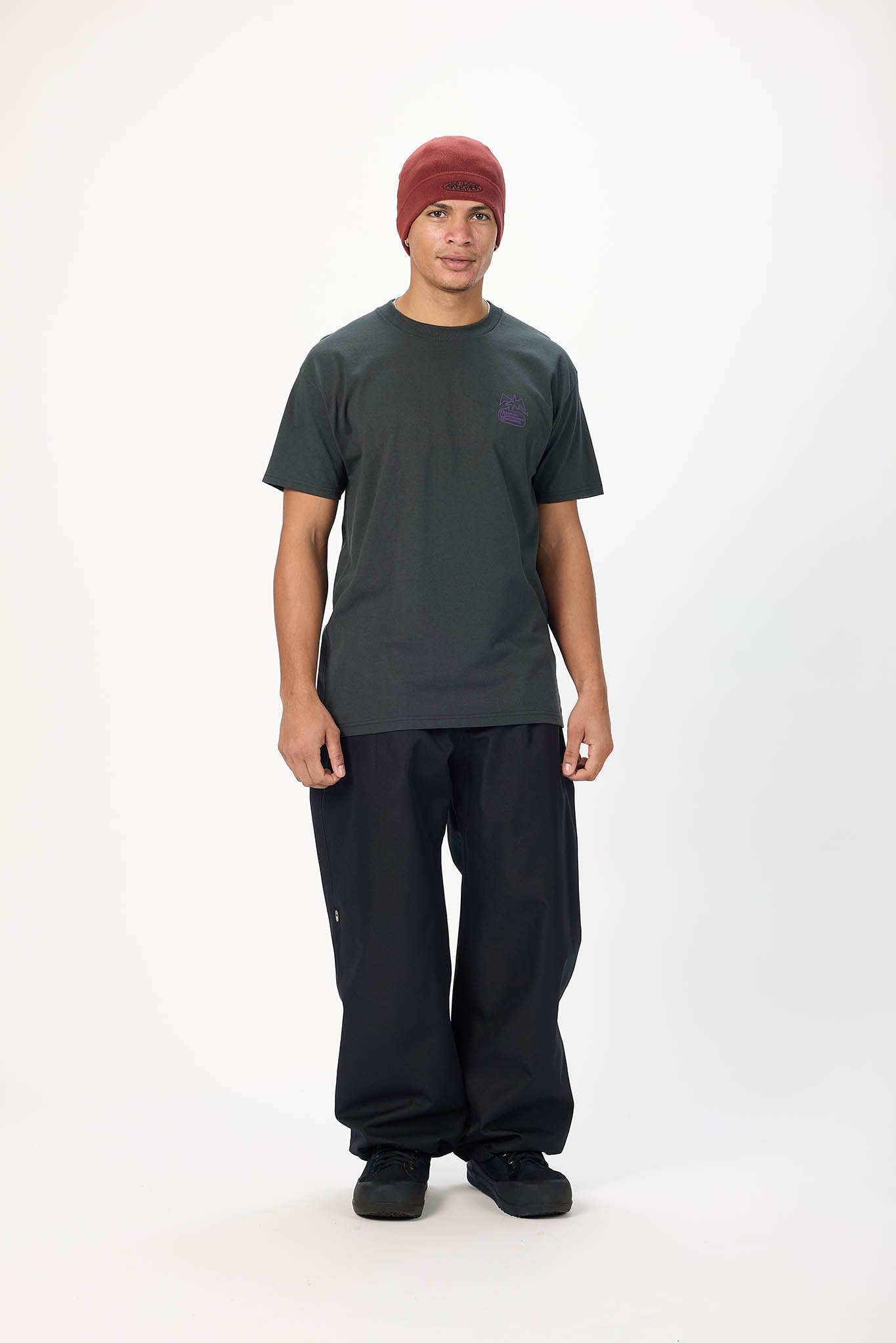 A man modeling a dark charcoal gray t-shirt with a small purple logo on the left chest, paired with black pants and a burgundy beanie, standing against a white studio background.