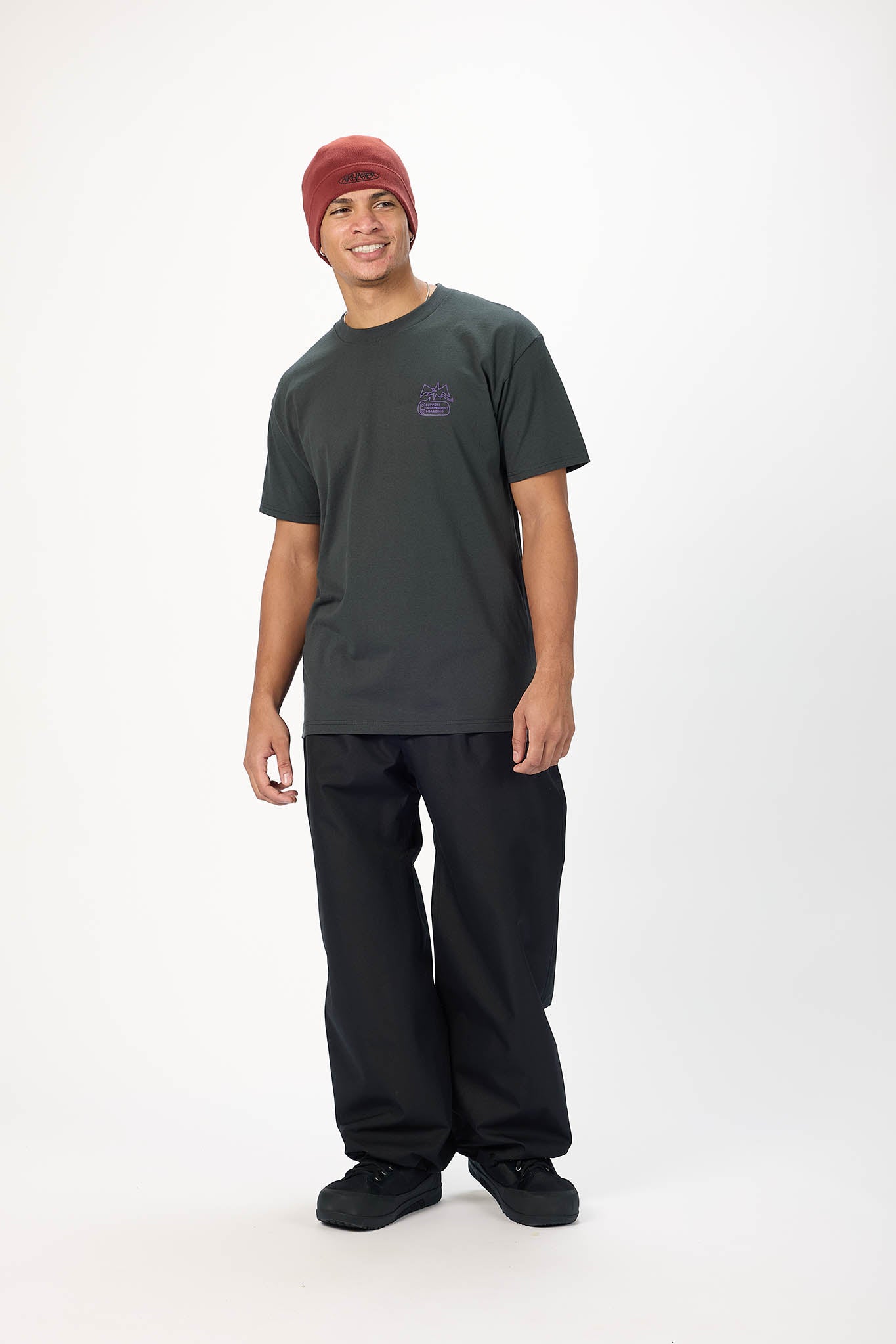 A man modeling a dark charcoal gray crewneck t-shirt with a small purple graphic on the left chest, paired with black pants, black shoes, and a rust-red beanie, standing against a plain white studio background.