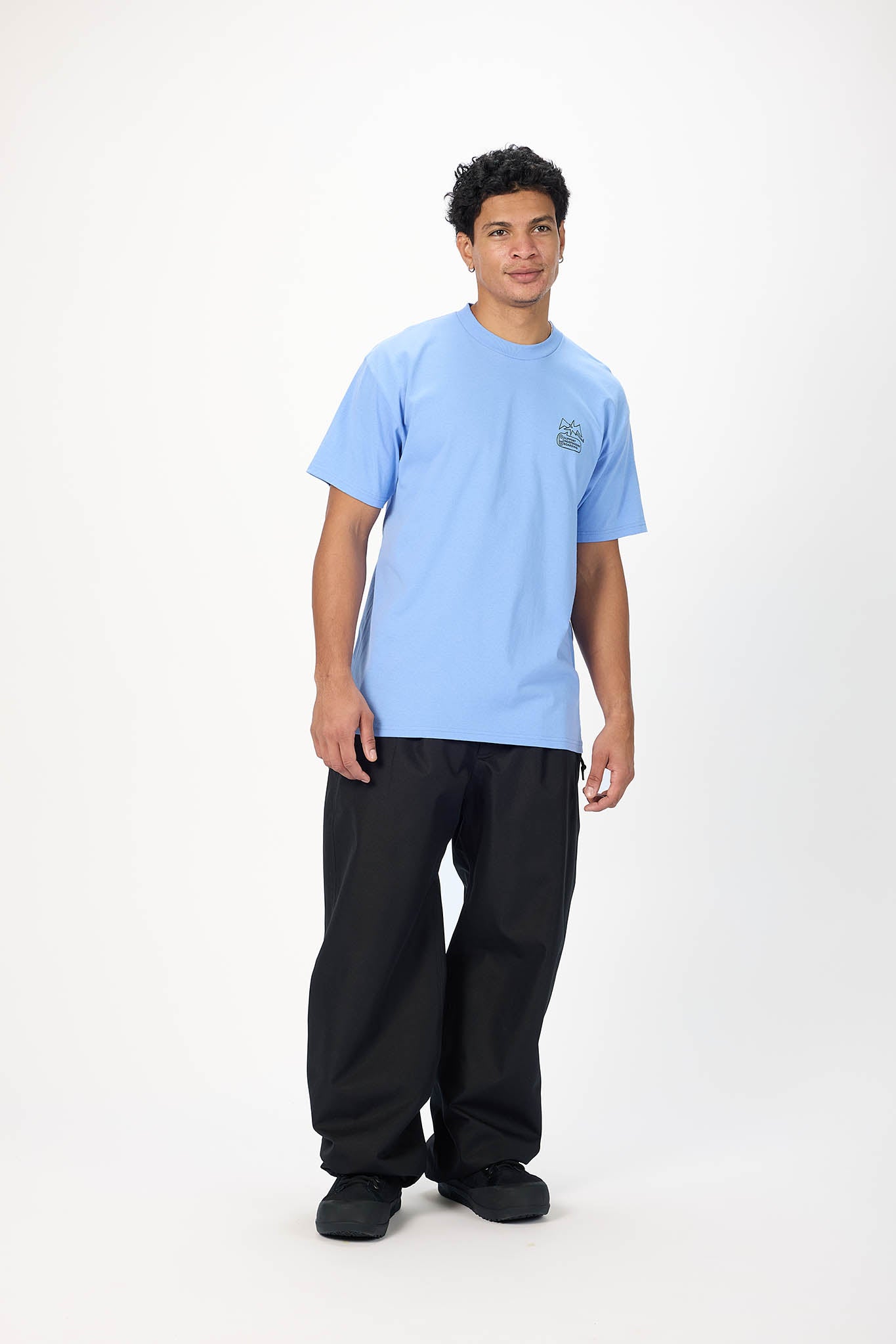 A man modeling a light blue crewneck t-shirt with a small green graphic on the left chest, paired with black pants, against a plain white studio background.