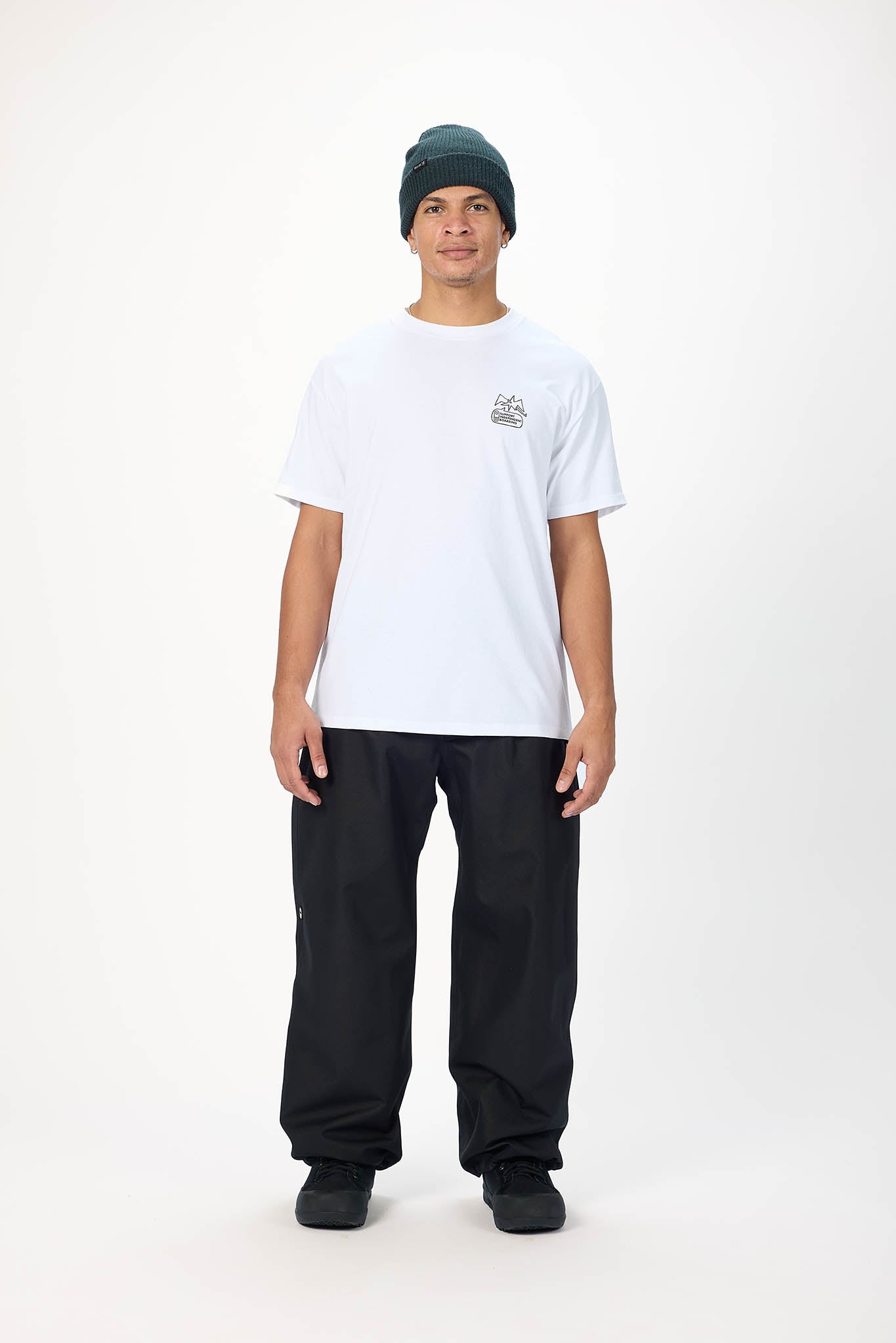 A model standing against a white studio background wearing a white crewneck t-shirt with a small chest graphic, black pants, and a dark green beanie.
