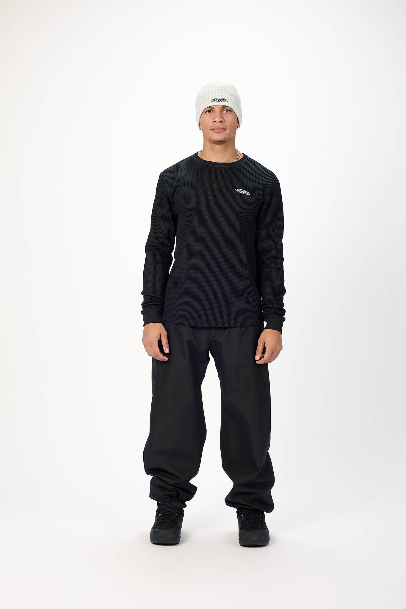 A man modeling a black long-sleeve crewneck sweatshirt, black pants, black boots, and a white beanie against a plain white studio background.