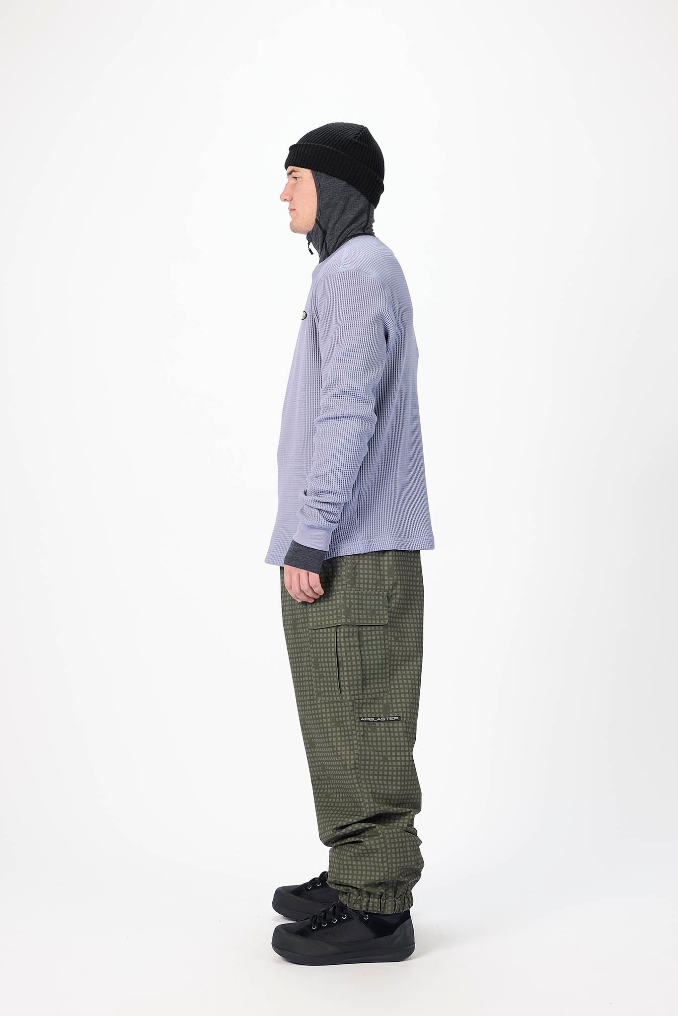 A model shown in profile wearing a light purple hooded jacket, olive green patterned pants, a black beanie, and black shoes, photographed against a plain white studio background.