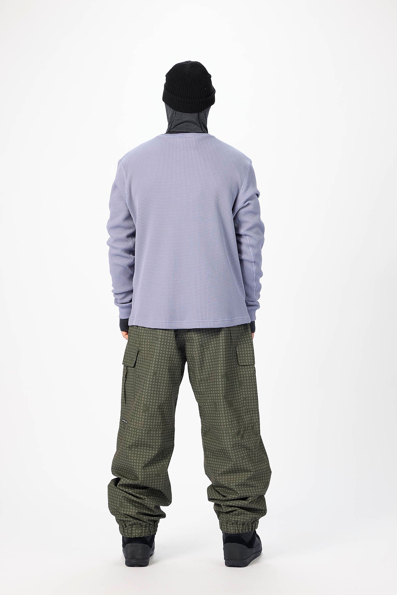 A model shown from the back wearing a light purple long-sleeve top, olive green patterned pants, a black beanie, and black shoes, against a plain white studio background.