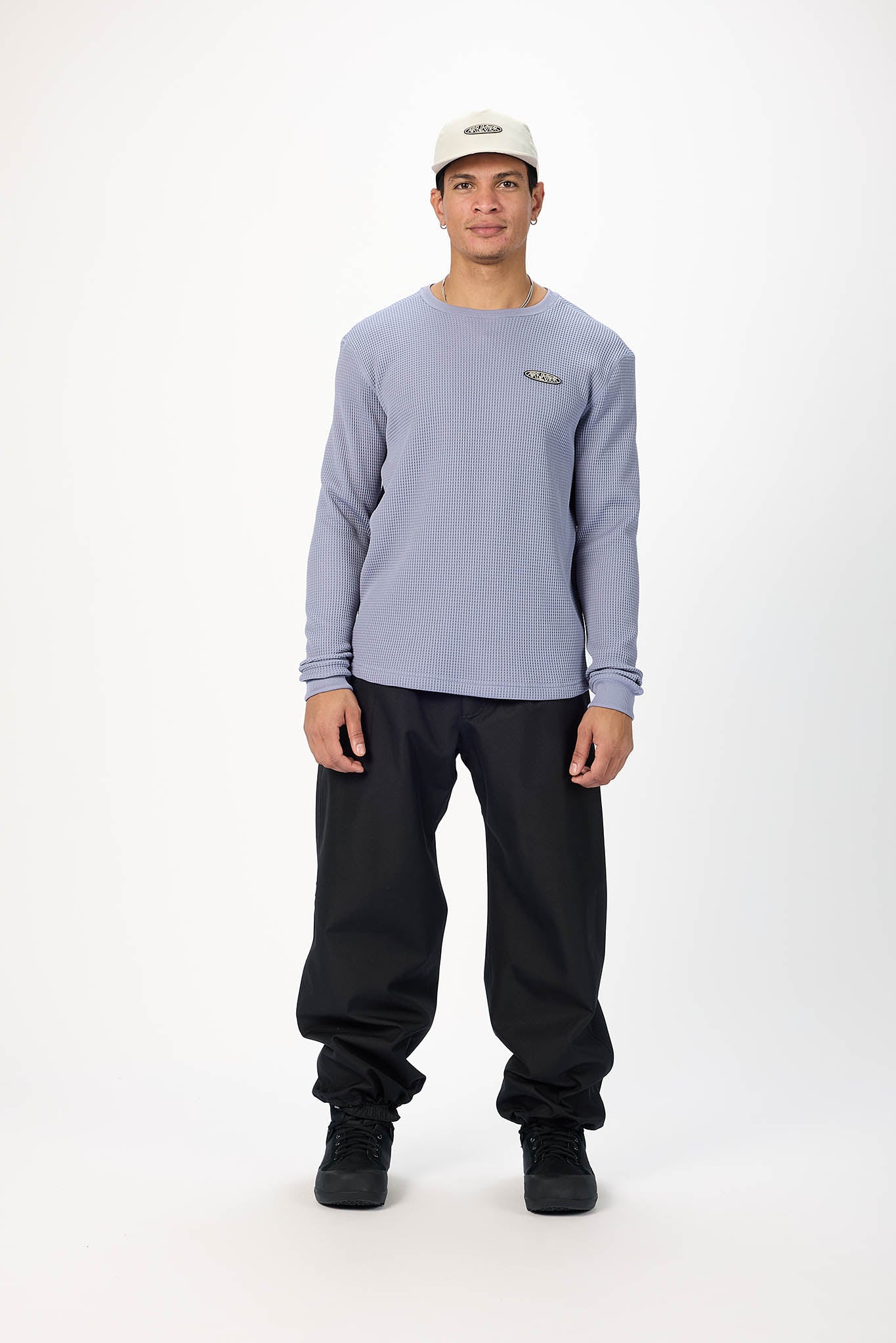 A man modeling a light periwinkle blue long-sleeve crewneck shirt with a small green crocodile logo on the left chest, paired with black sweatpants, black sneakers, and a white cap, against a plain white studio background.