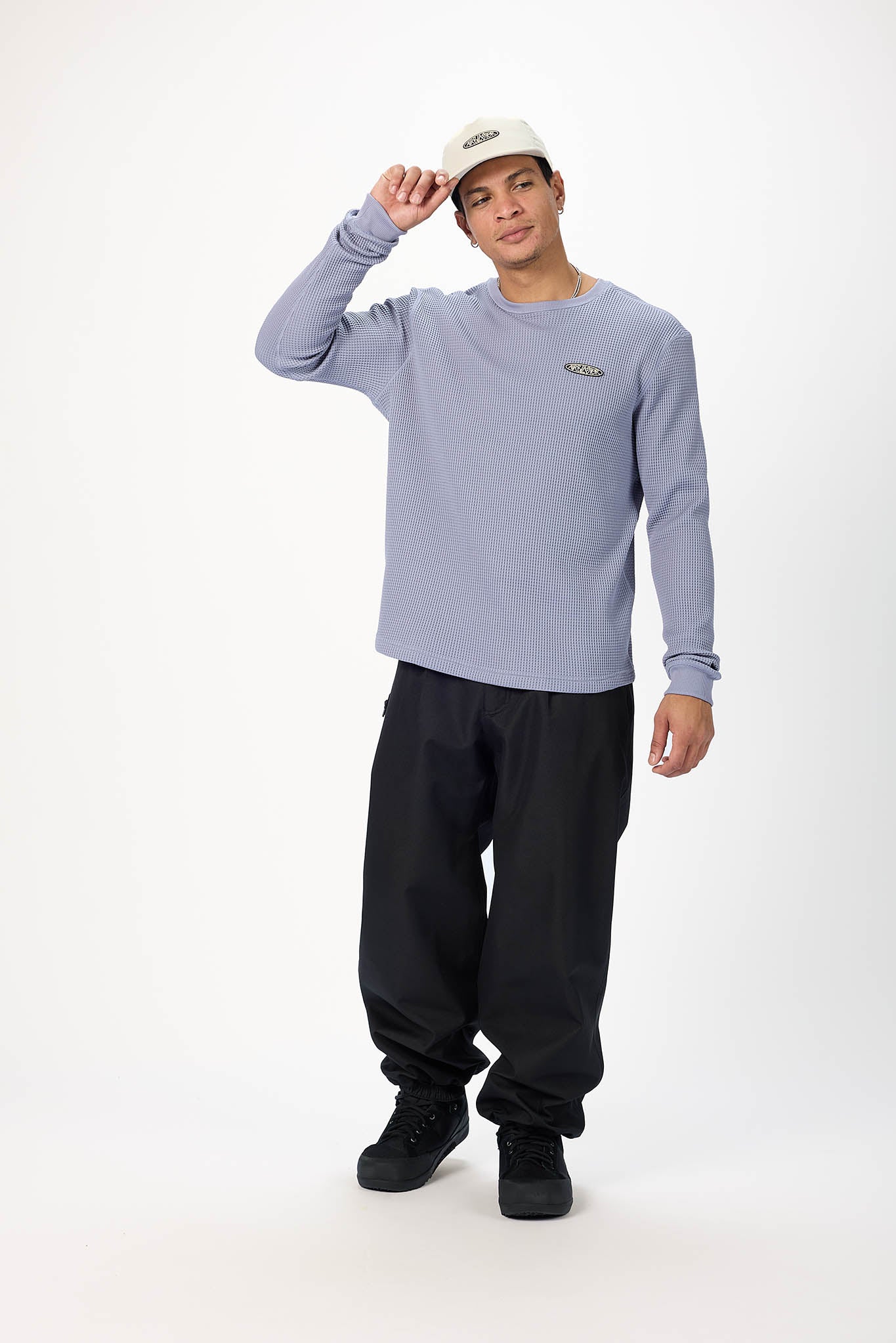 A man modeling a light periwinkle blue long-sleeve crewneck shirt, black sweatpants, black sneakers, and a white baseball cap against a plain white studio background.