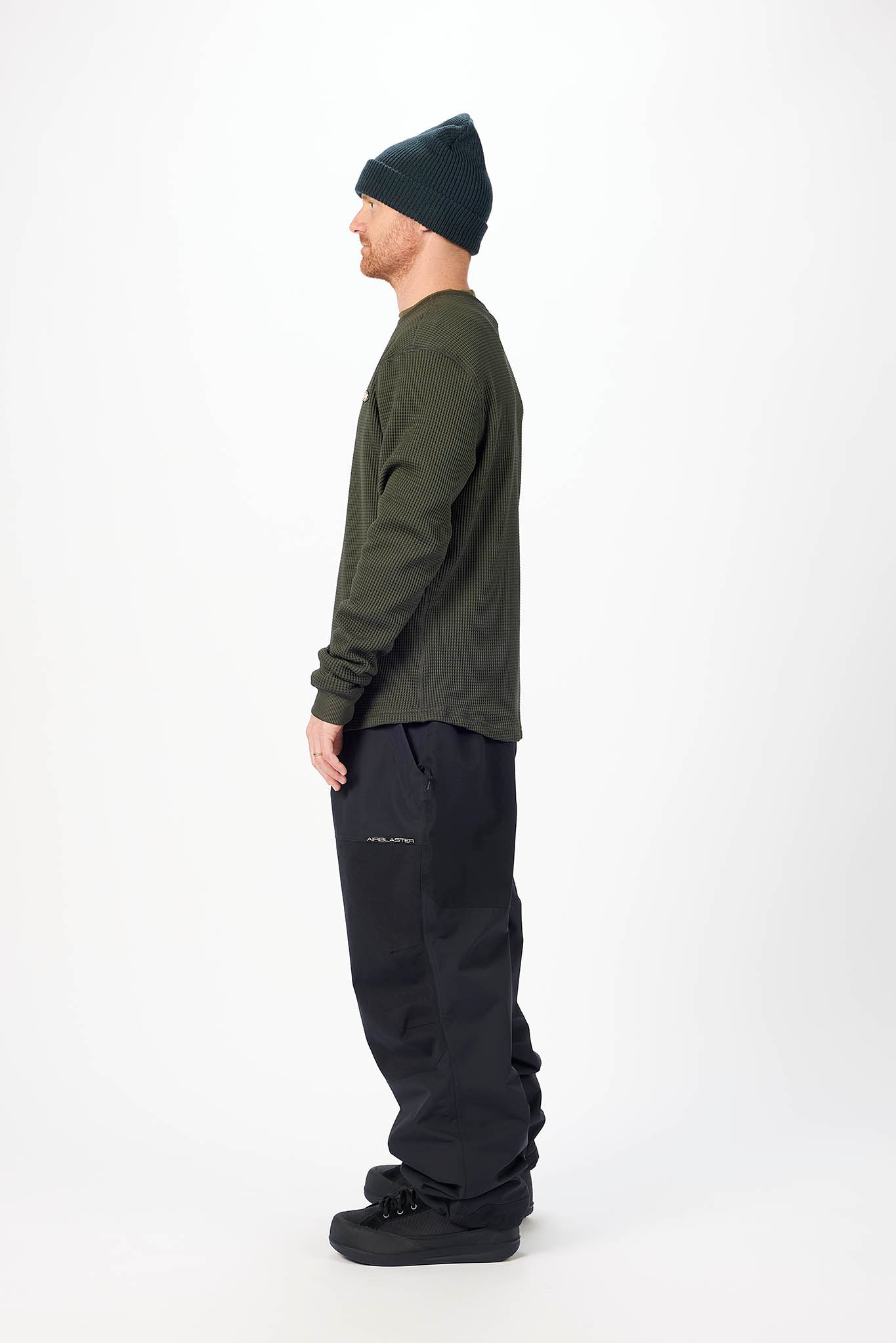 A man shown in profile wearing a dark olive green long-sleeve shirt, black pants, a black beanie, and black shoes, standing against a plain white studio background.