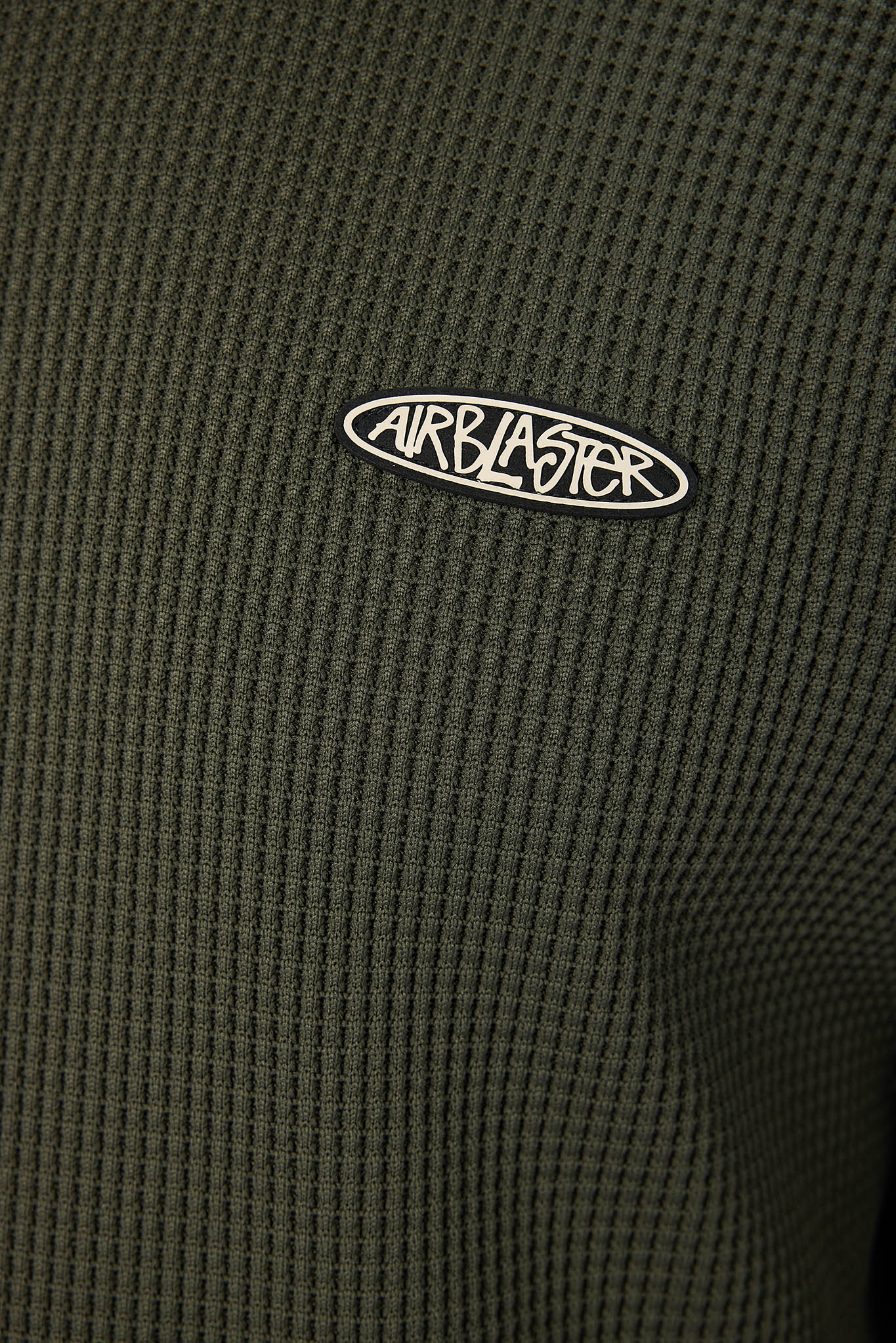 Close-up of a dark olive green waffle-knit fabric with a white and black oval Airblaster logo patch on the chest.