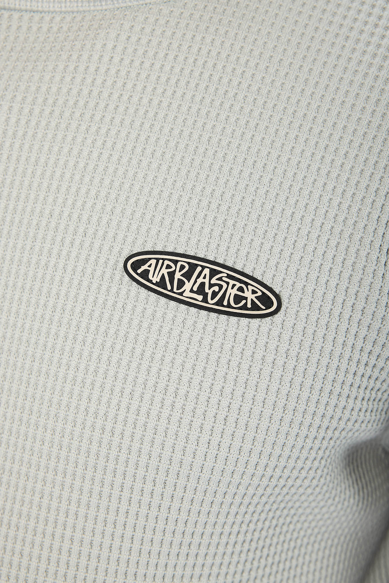 Close-up of a light gray waffle-knit sweatshirt featuring a black and white oval Airblaster logo patch on the chest.