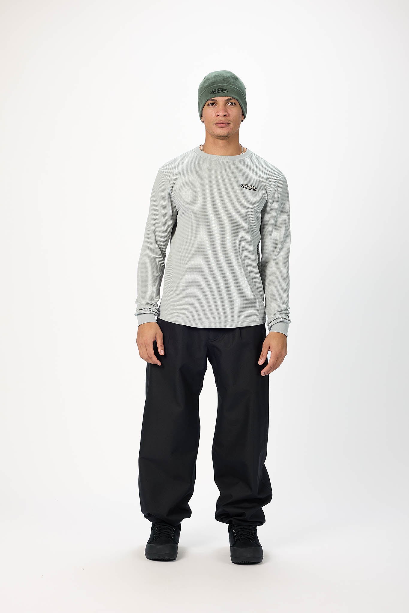 A man modeling a light gray long-sleeve crewneck sweatshirt, black pants, and a green beanie against a plain white studio background.