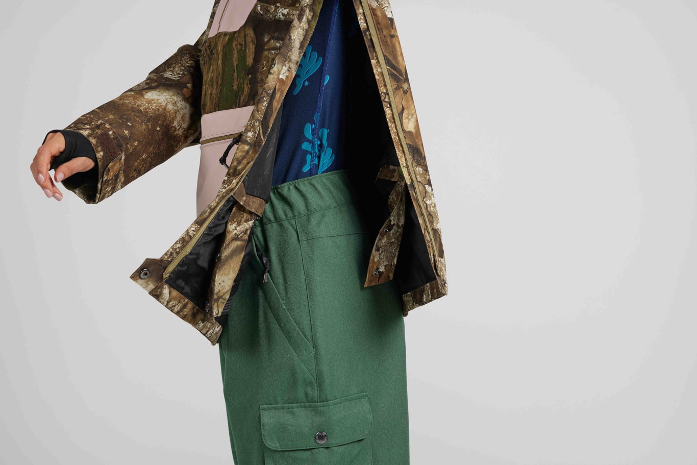 A person wearing a brown and tan camouflage jacket over a pink and blue top, paired with olive green cargo pants, shown against a light gray studio background.