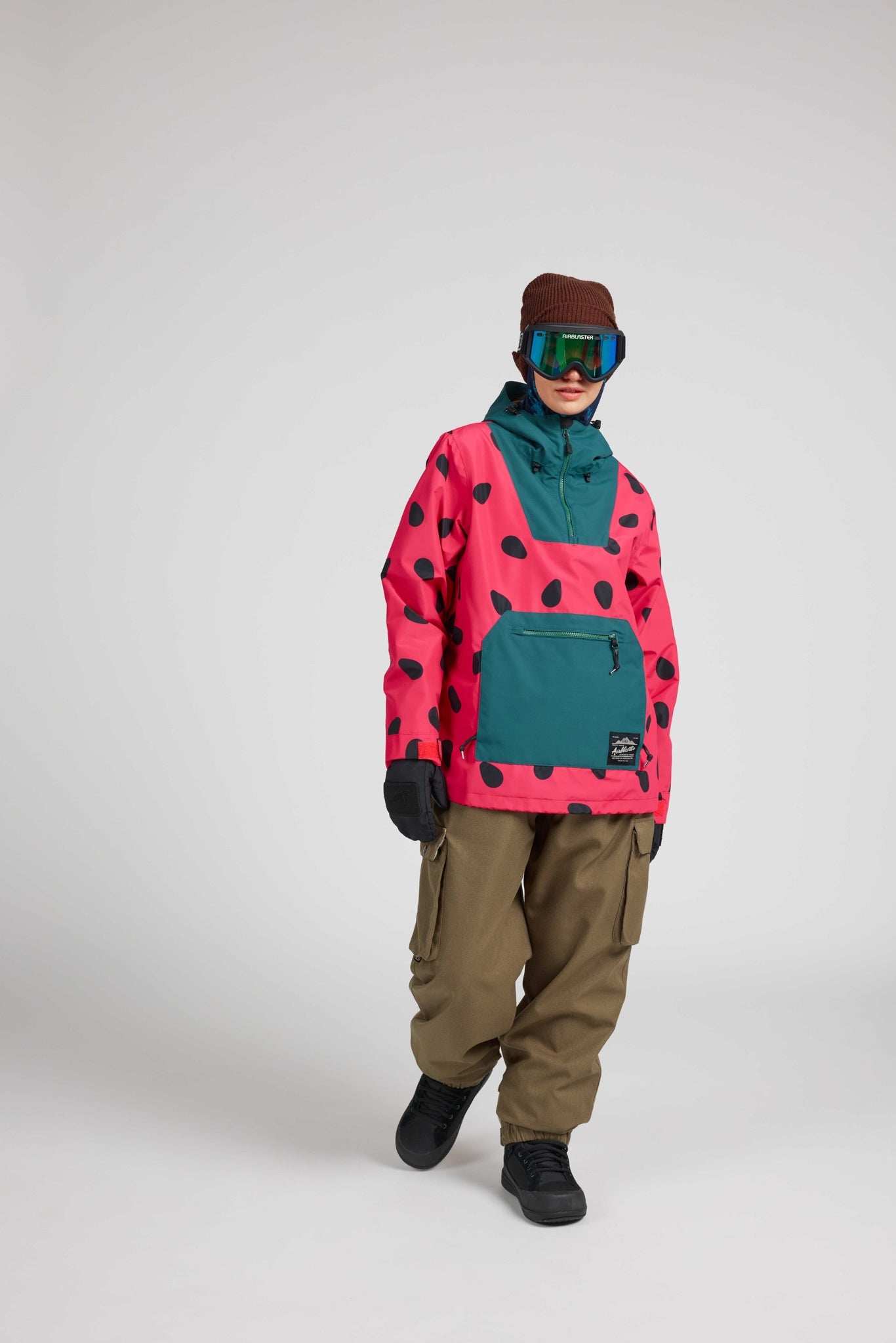 A model wearing a pink and teal snowboard jacket with black spots, brown pants, a brown beanie, and mirrored goggles, standing against a light gray studio background.